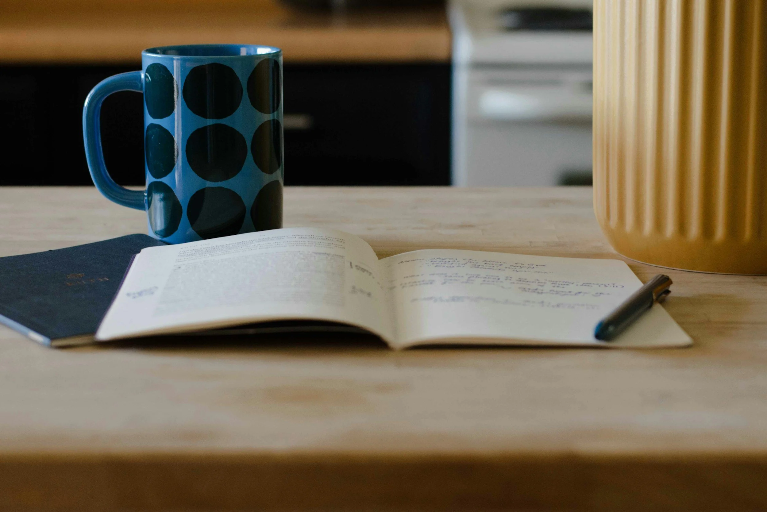Open notebook with handwritten notes, a pen, a blue mug with black polka dots, and a yellow vase on a wooden table in a kitchen or dining area.