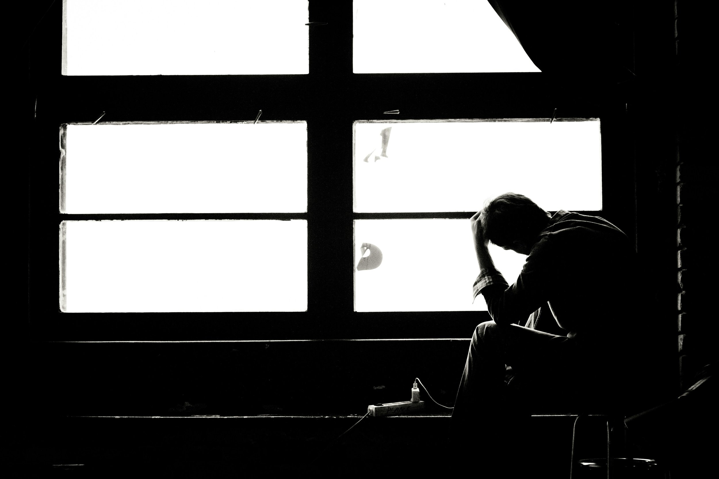 Black and white image of man sitting and holding his head looking out a window