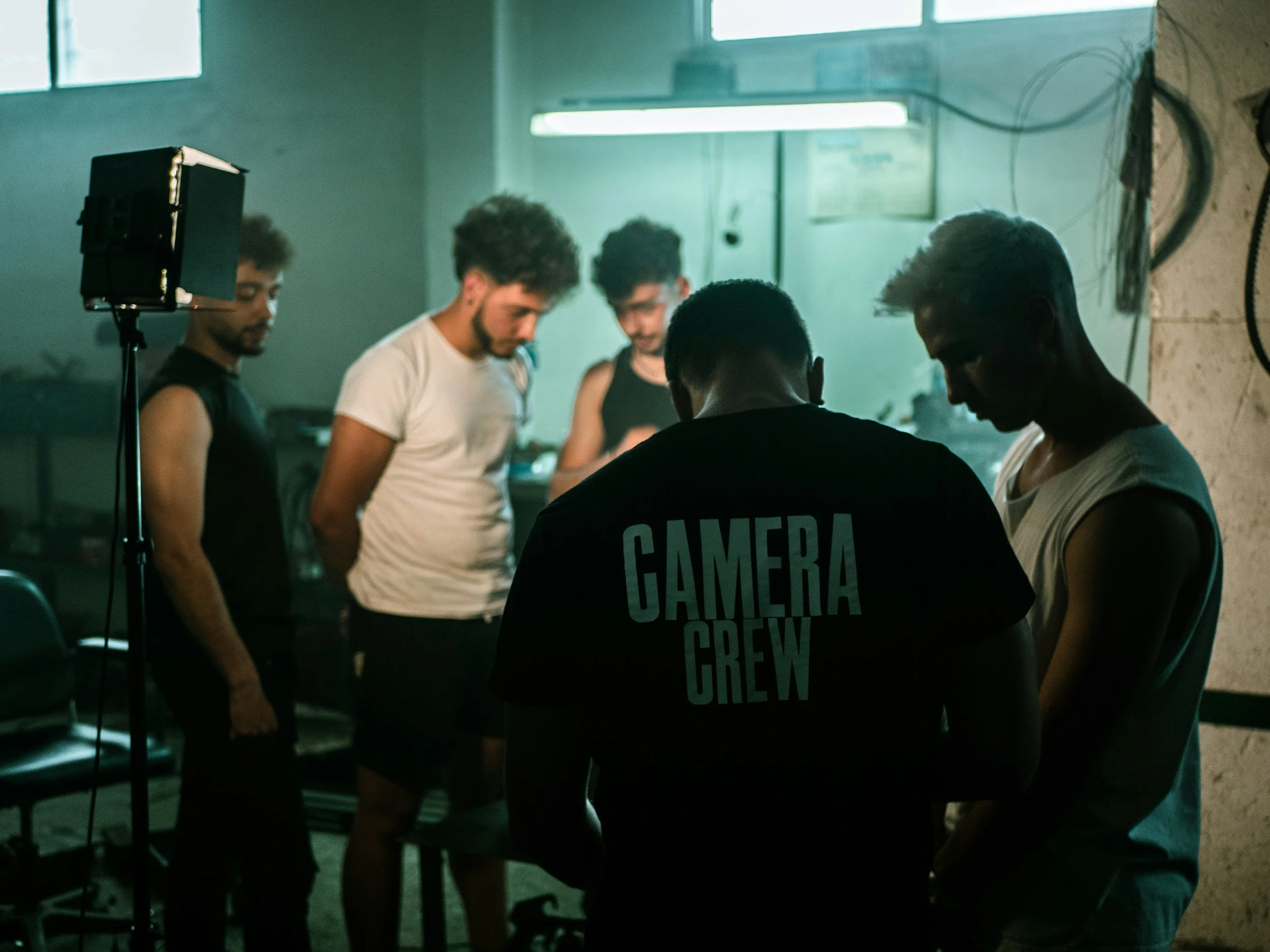 Five people in a dimly lit room, with one wearing a shirt labeled "Camera Crew." They appear to be engaged in a discussion or preparation for filming, with filming equipment visible.
