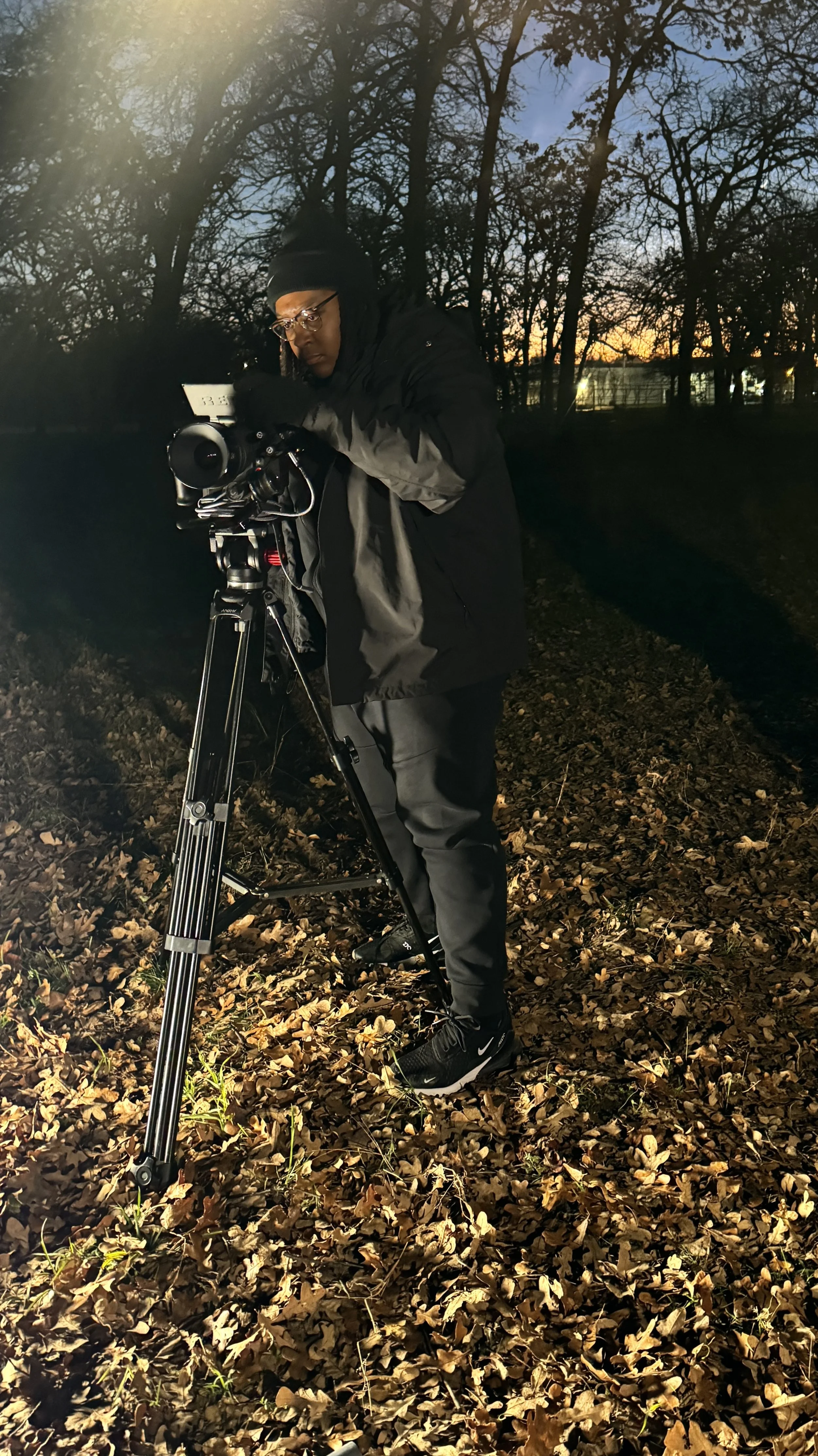 A person dressed warmly operates a camera on a tripod in a forest at dusk, with fallen leaves on the ground.