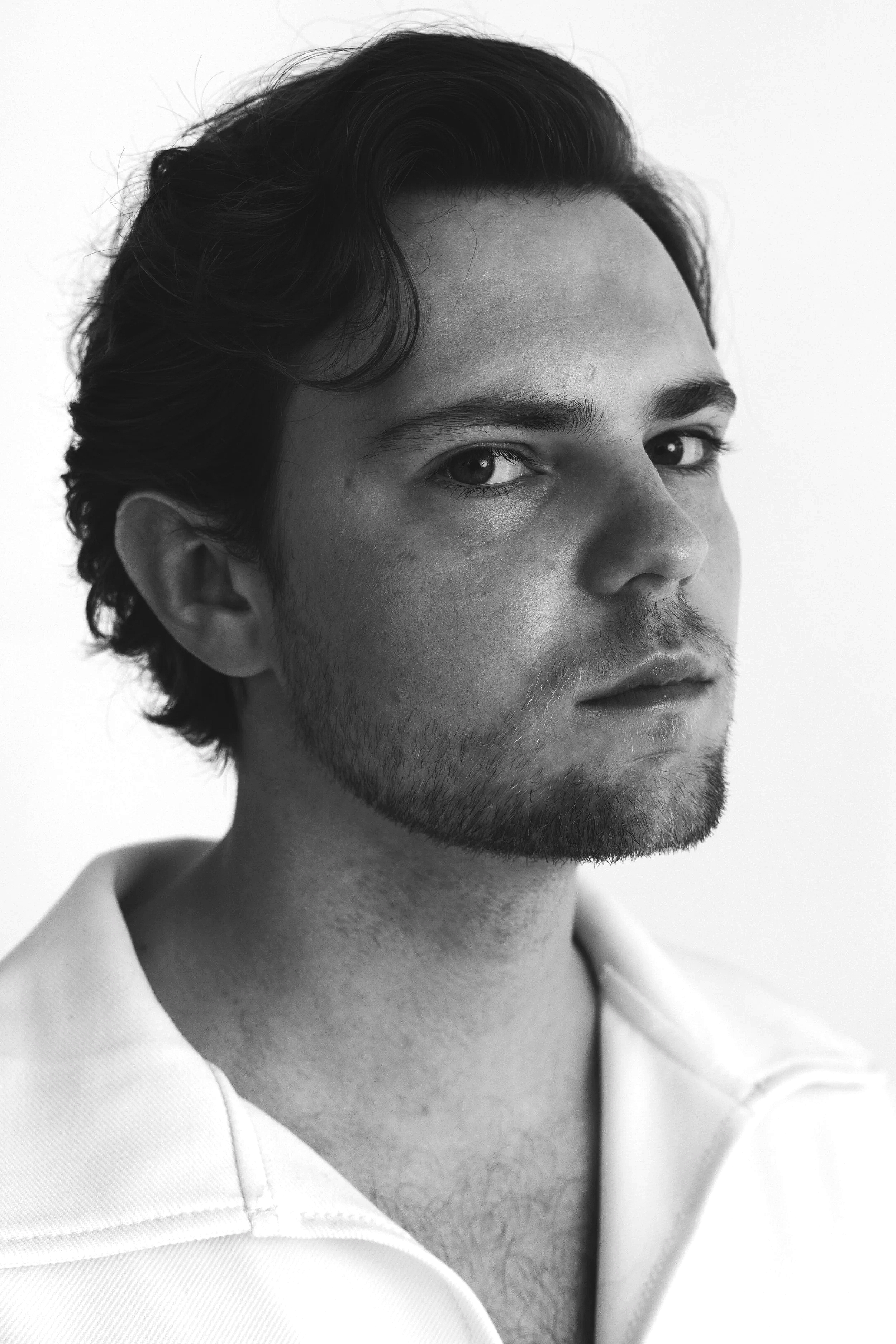 Close-up black and white portrait of a young man with wavy hair, light skin, and slight facial hair, wearing a collared shirt, looking slightly to the side.