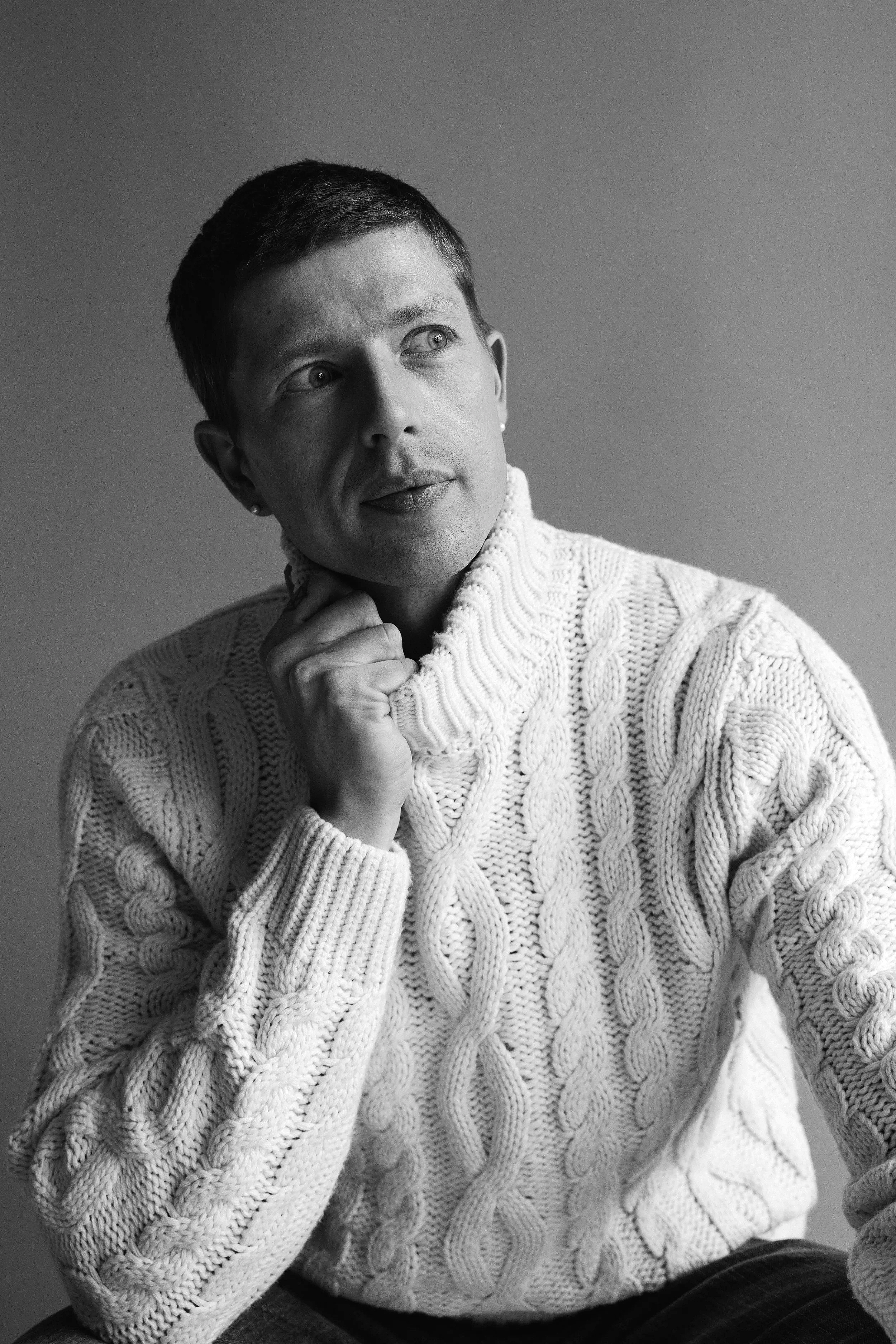 Black and white photo of a woman with short hair wearing a chunky knit sweater. She is looking to her left with a thoughtful expression, touching her chin with her right hand.