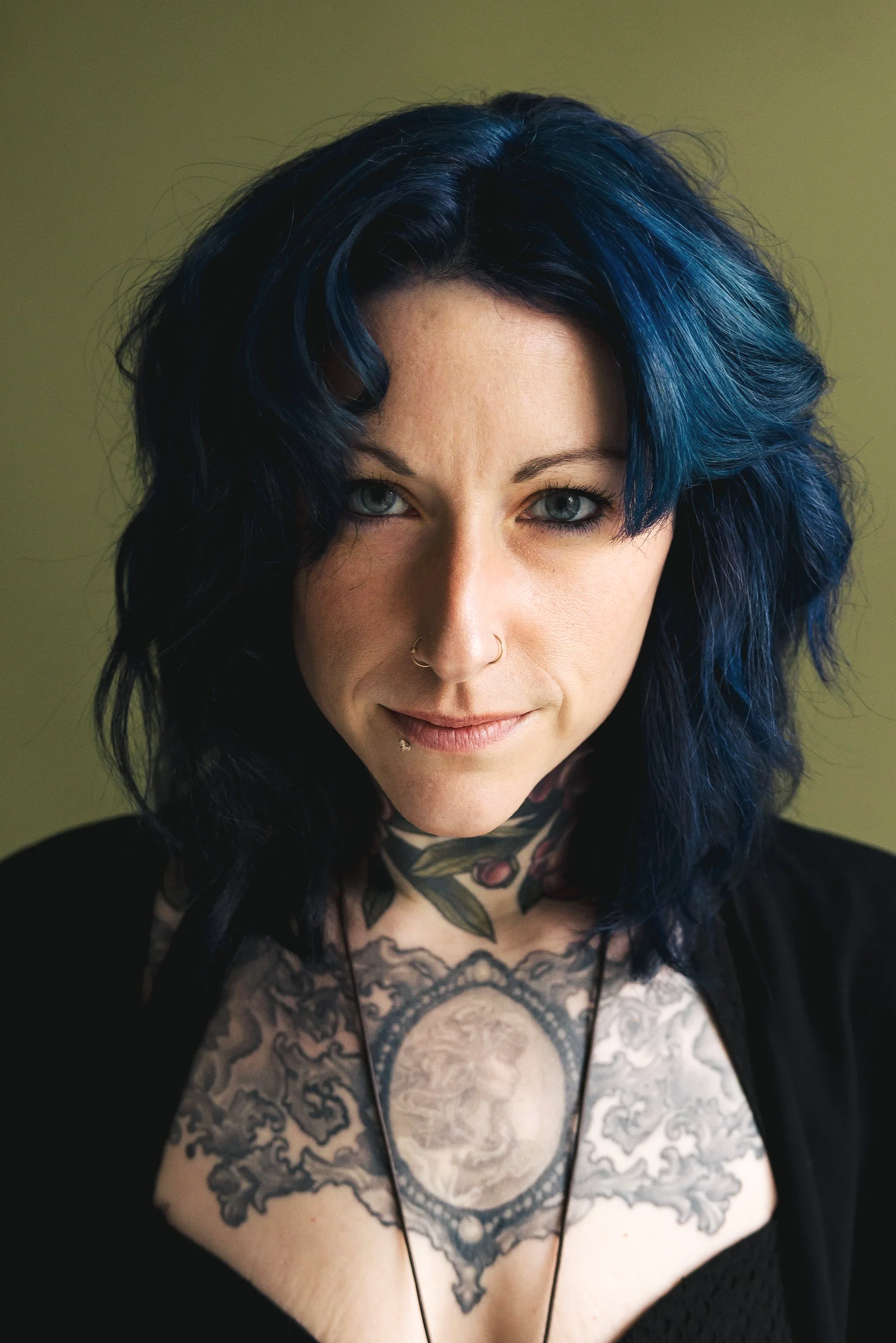 A woman with dark blue hair, blue eyes, and tattoos on her chest and shoulders, wearing a black top and a necklace, looking directly at the camera.