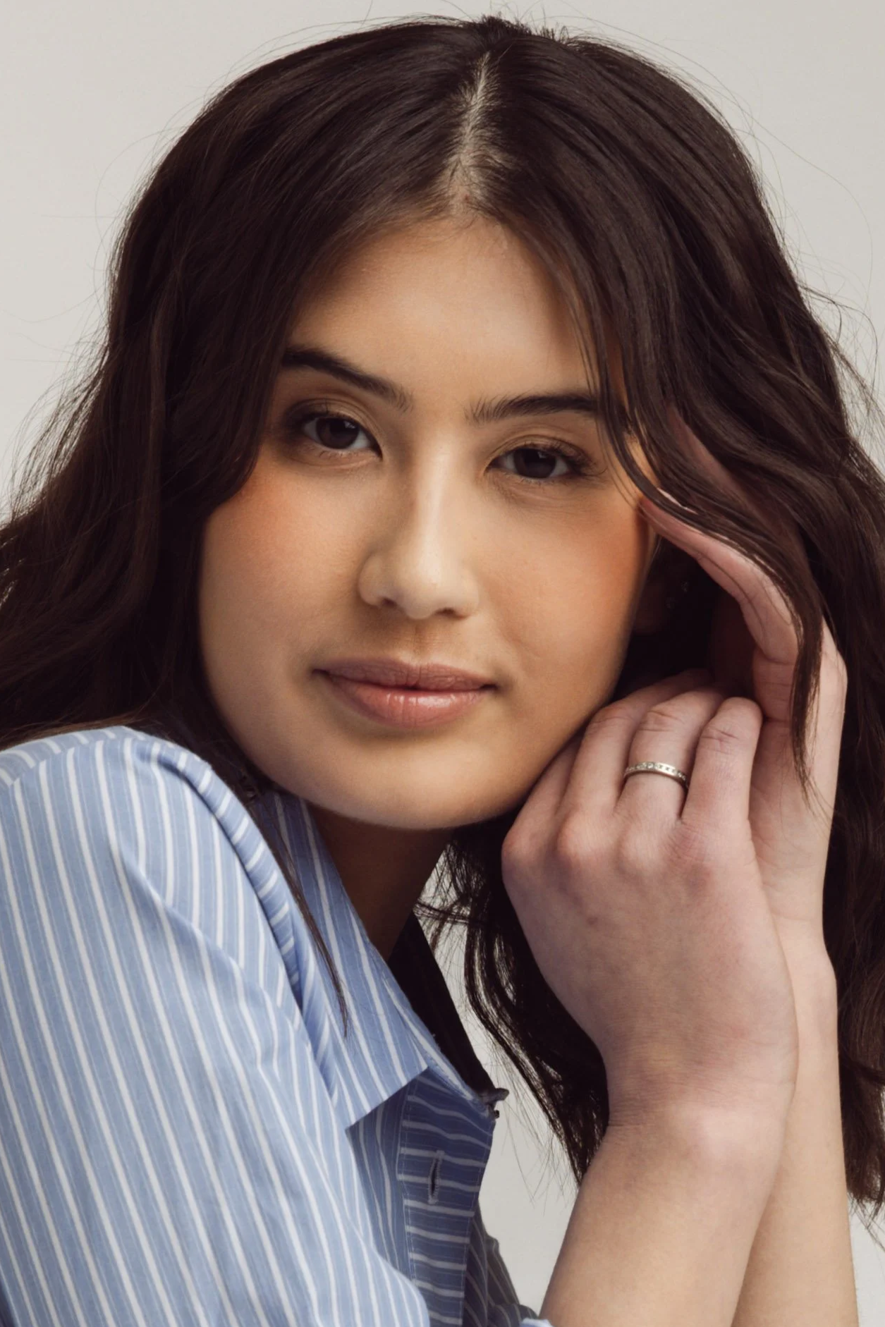 Close-up portrait of a woman with long brown hair, wearing a blue and white striped shirt, resting her head on her hand, and looking at the camera with a gentle expression.