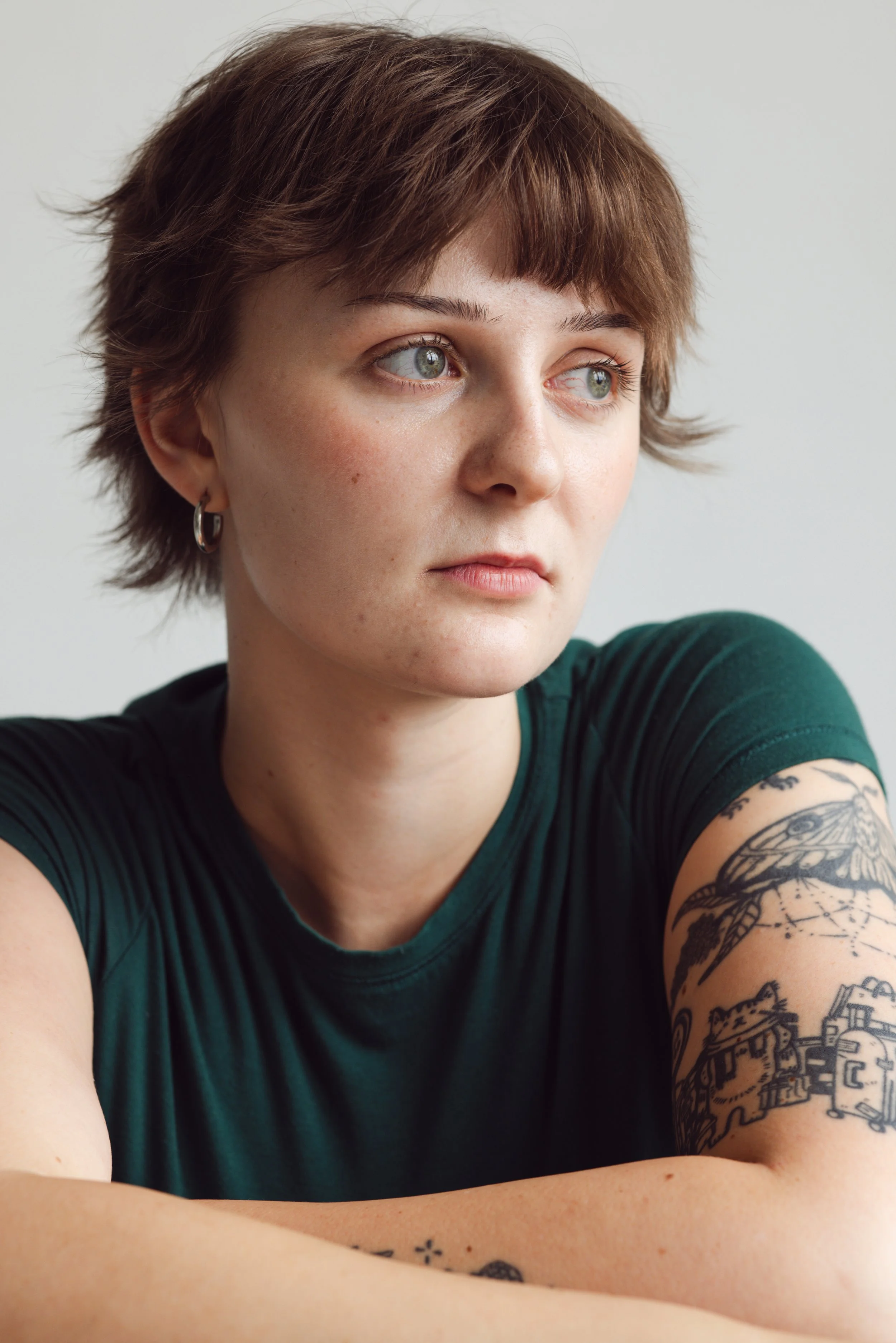Close-up portrait of a young woman with short brown hair and green eyes, wearing a dark green t-shirt and a silver hoop earring, displaying a tattoo sleeve on her right arm, and looking thoughtfully off to the side.