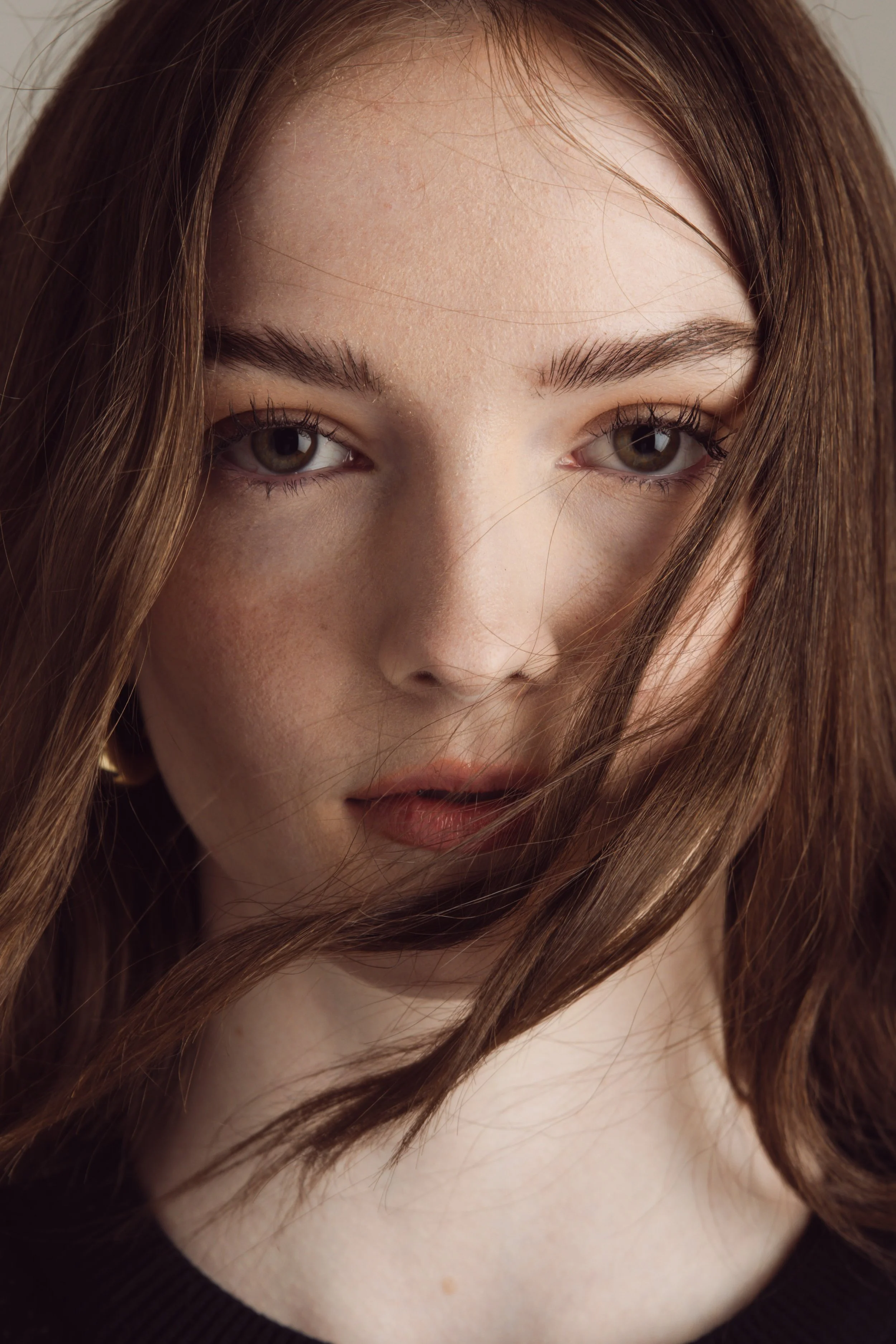 Close-up of a woman's face with brown hair partially covering her face, hazel eyes, and wearing a dark top.