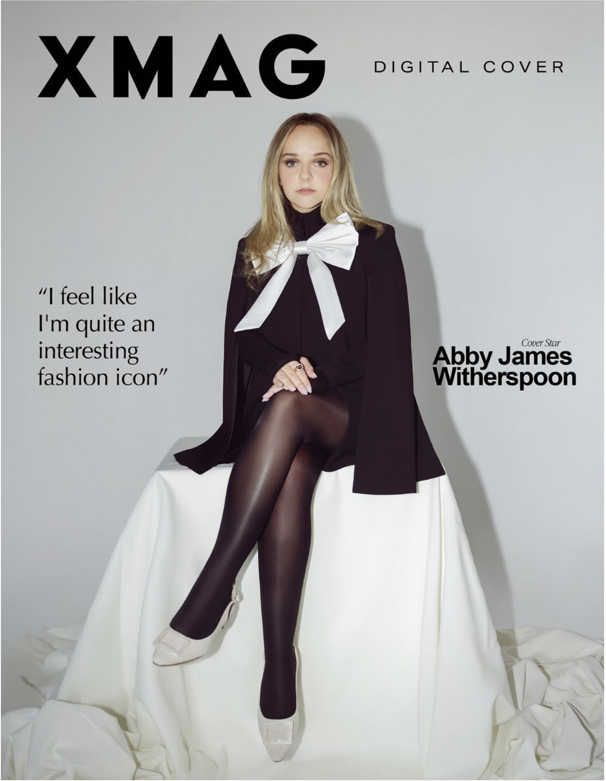 Cover of XMAG magazine featuring Abby James Witherspoon sitting on a white draped surface, wearing a black blazer, sheer tights, white heels, and a large white bow tie, with a quote and magazine title.