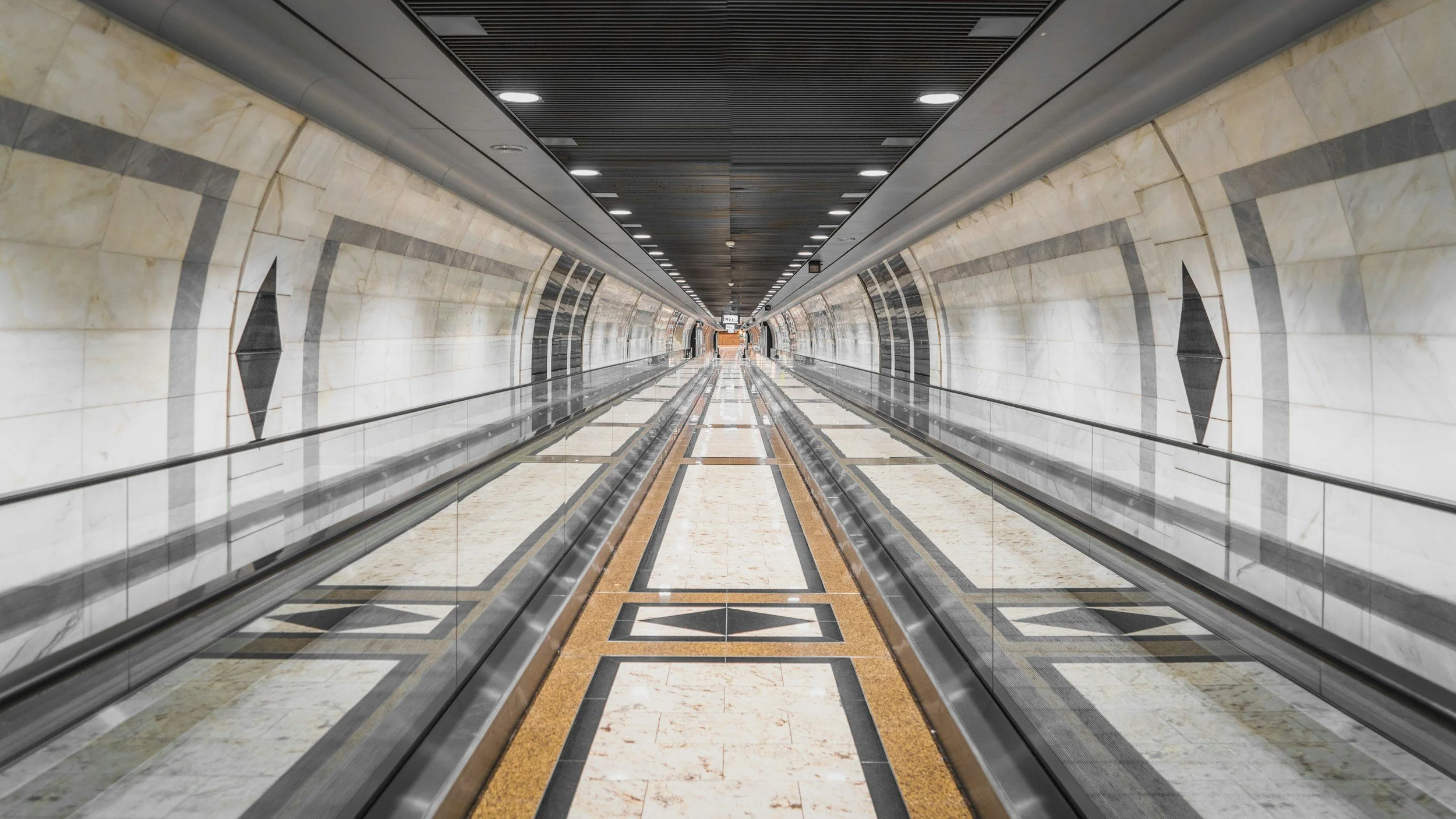 Long underground tunnel with moving walkways and marble walls, ceiling with circular recessed lights