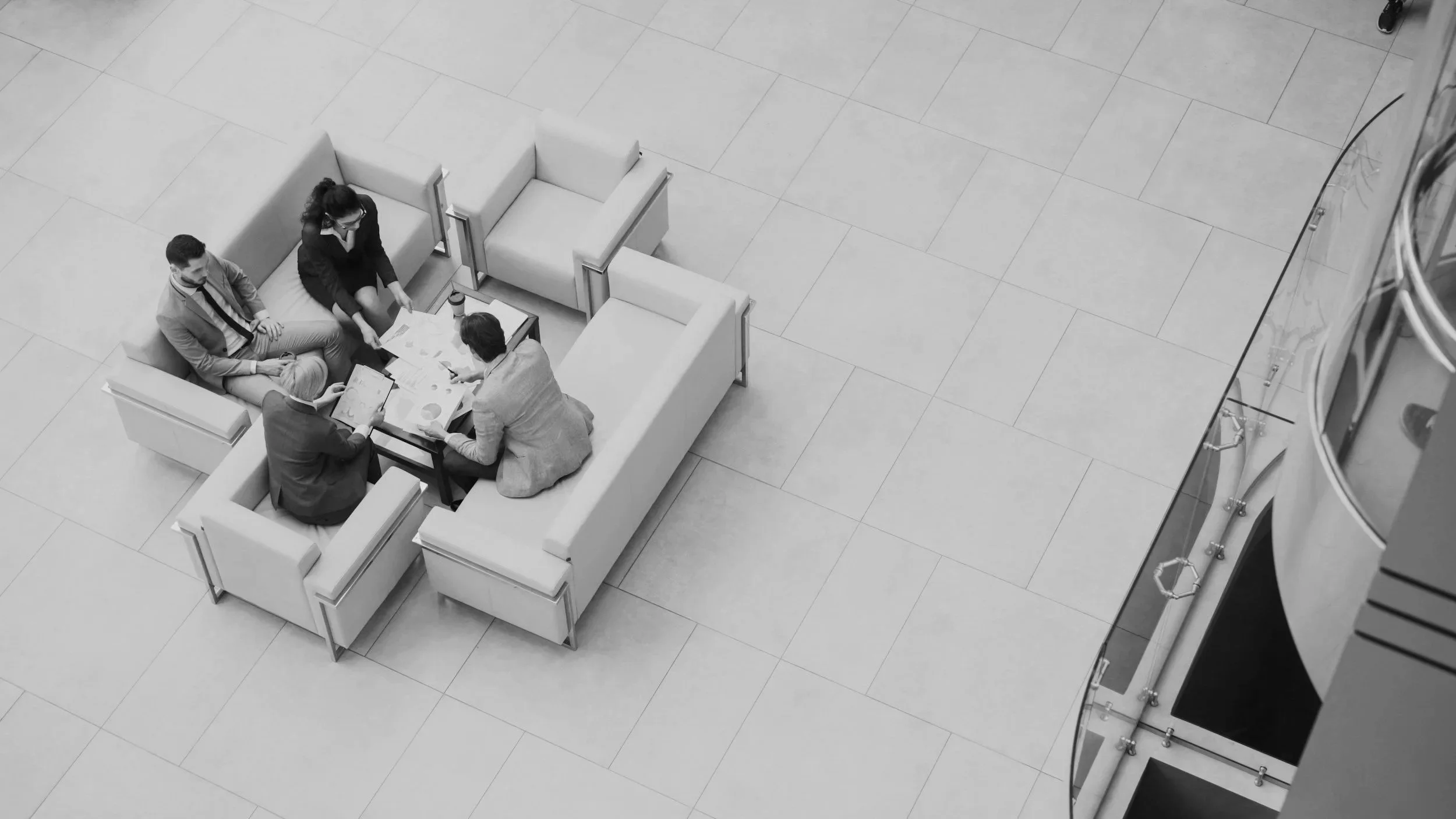 A group of five people are sitting around a table having a meeting in a lobby with light-colored tiled floor and some couches.