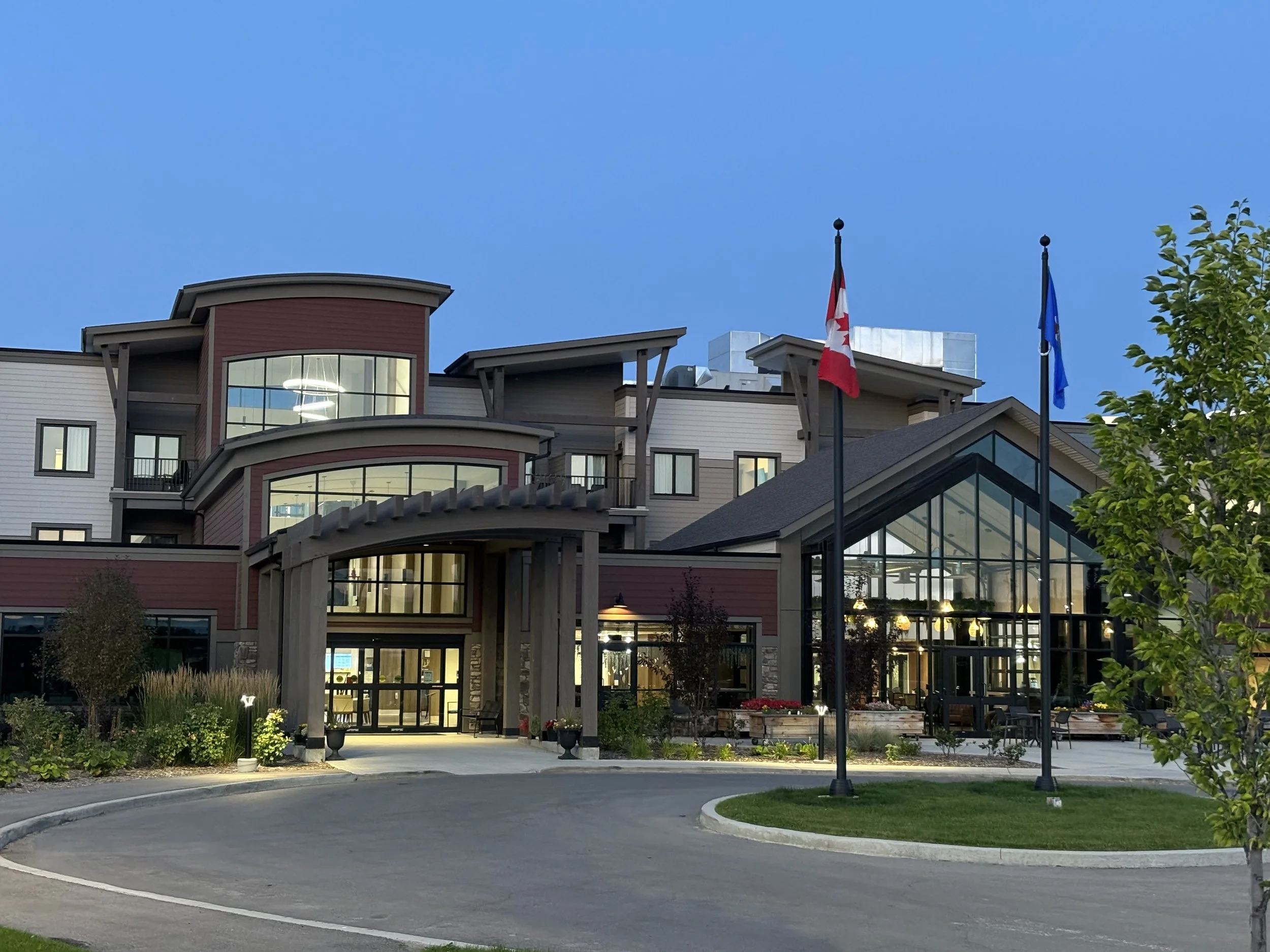 Exterior view of a modern multi-story building with large glass windows, featuring a red and gray color scheme. Two flags, one Canadian and one blue, are flying on flagpoles in front. The building is surrounded by landscaped greenery and a circular d