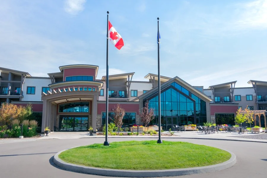 Modern multi-story building with large glass windows and outdoor seating, featuring the Canadian flag and another flag on poles in front, surrounded by a lawn and trees.