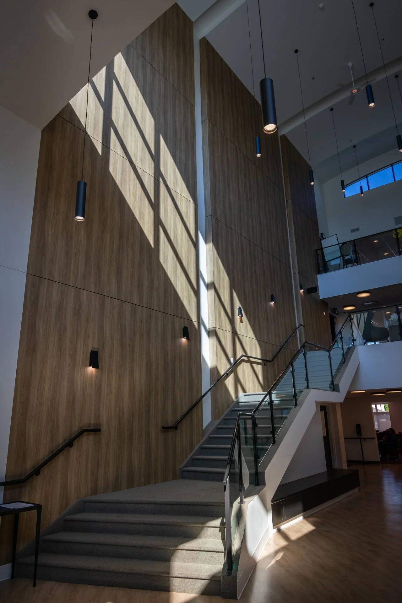 Modern interior with high wooden wall, staircase with glass railing, and pendant lights casting shadows on the wall.