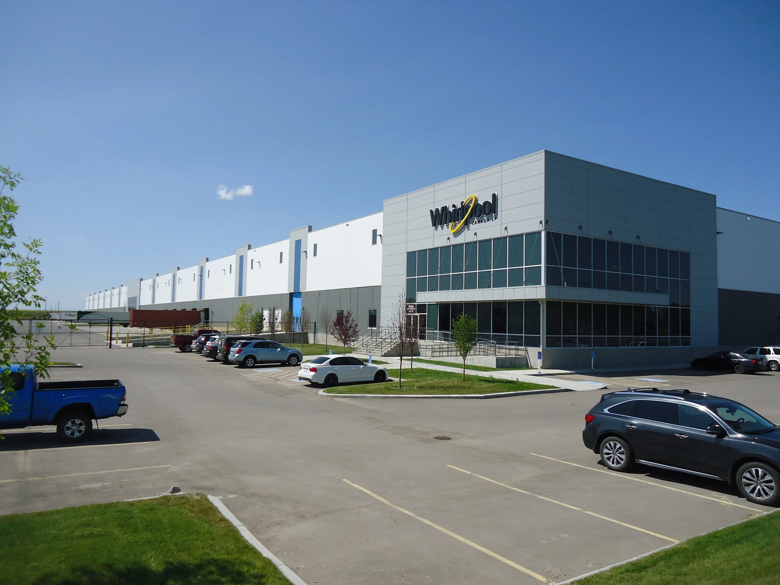 Large commercial building with a sign reading 'Whirlpools Canada' and a parking lot with several parked cars, under a clear blue sky.