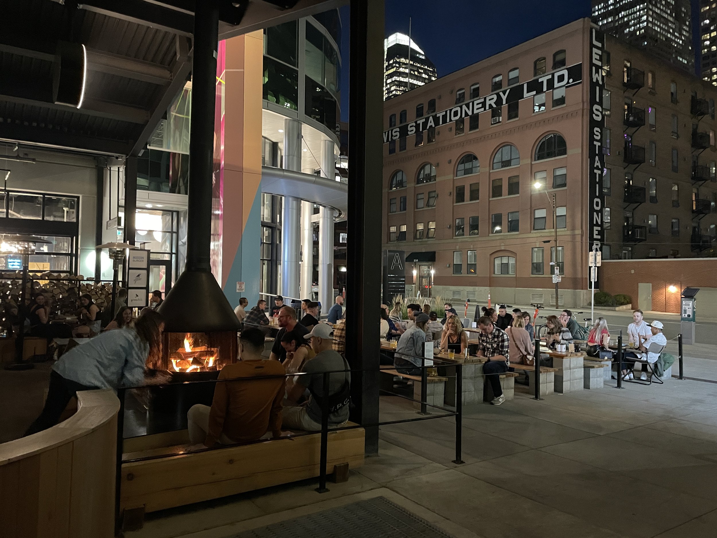 People dining outside at night with string lights, a fireplace, and a cityscape background.