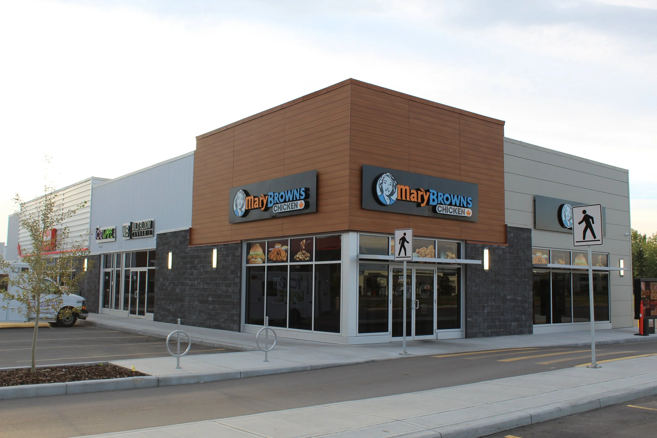 A shopping plaza with stores including Mary Browns Chicken, with signs for other stores like Wild Bloom Wellness and Vape. The building has a modern design with sidewalk signs, trees, and parking spaces in the foreground.