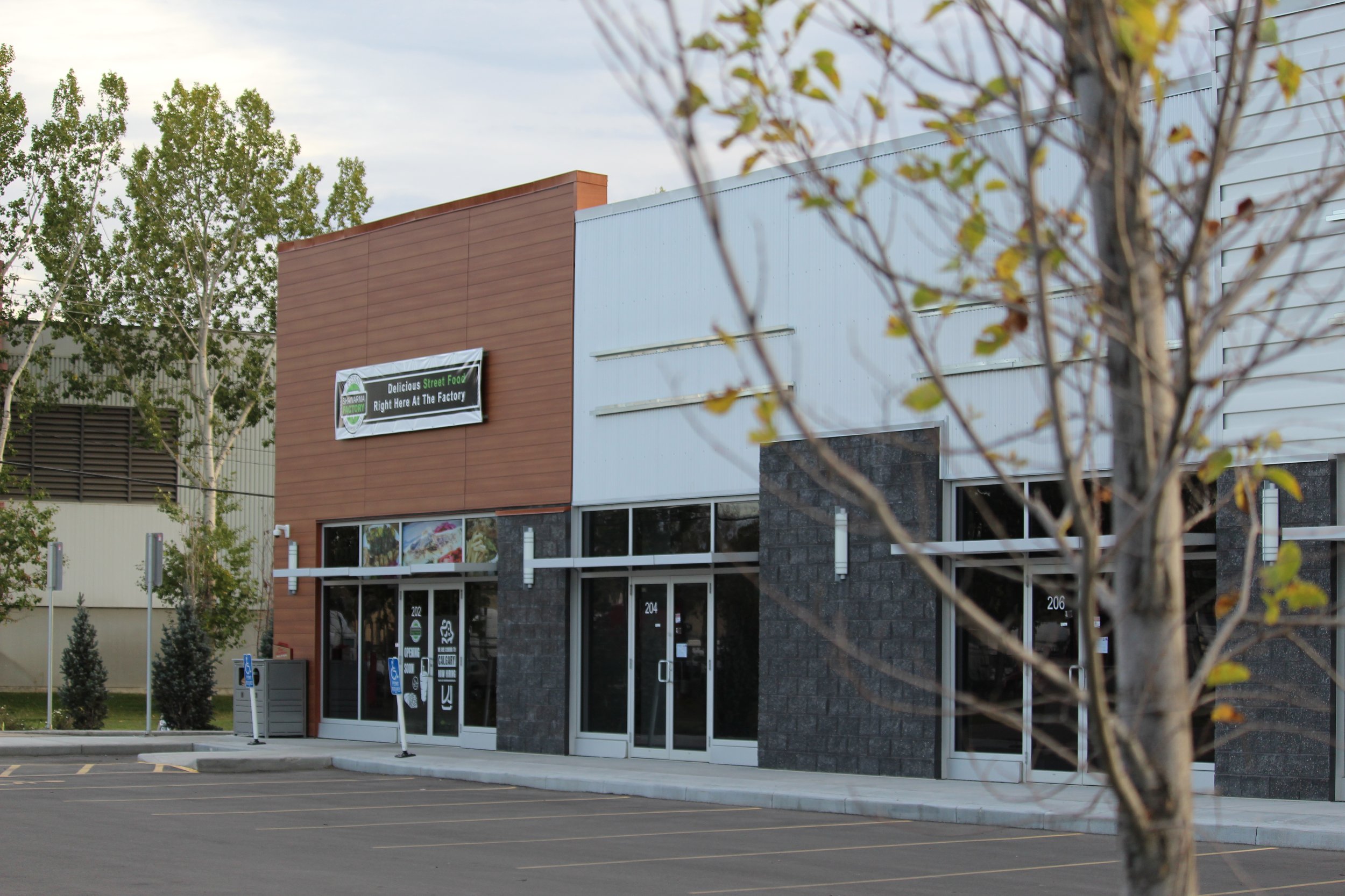 Exterior view of a modern commercial building with a sign indicating street food options, a parking lot in the foreground, and trees nearby.