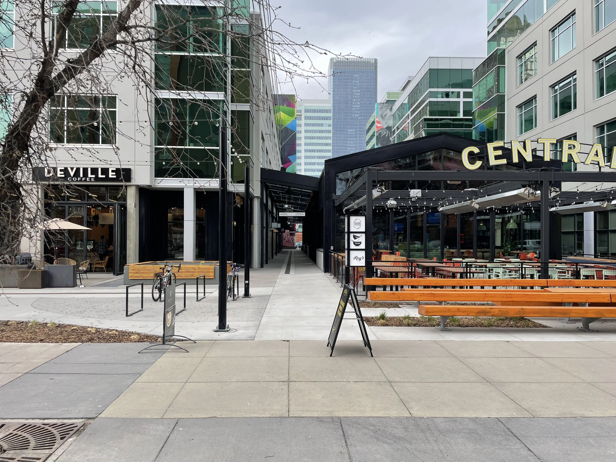 A modern city plaza with a sidewalk, benches, and trees, surrounded by multi-story glass buildings; a coffee shop named DEVILLE and an outdoor dining area with orange seating, string lights, and a black structure with yellow sign spelling 'CENTRAL'.