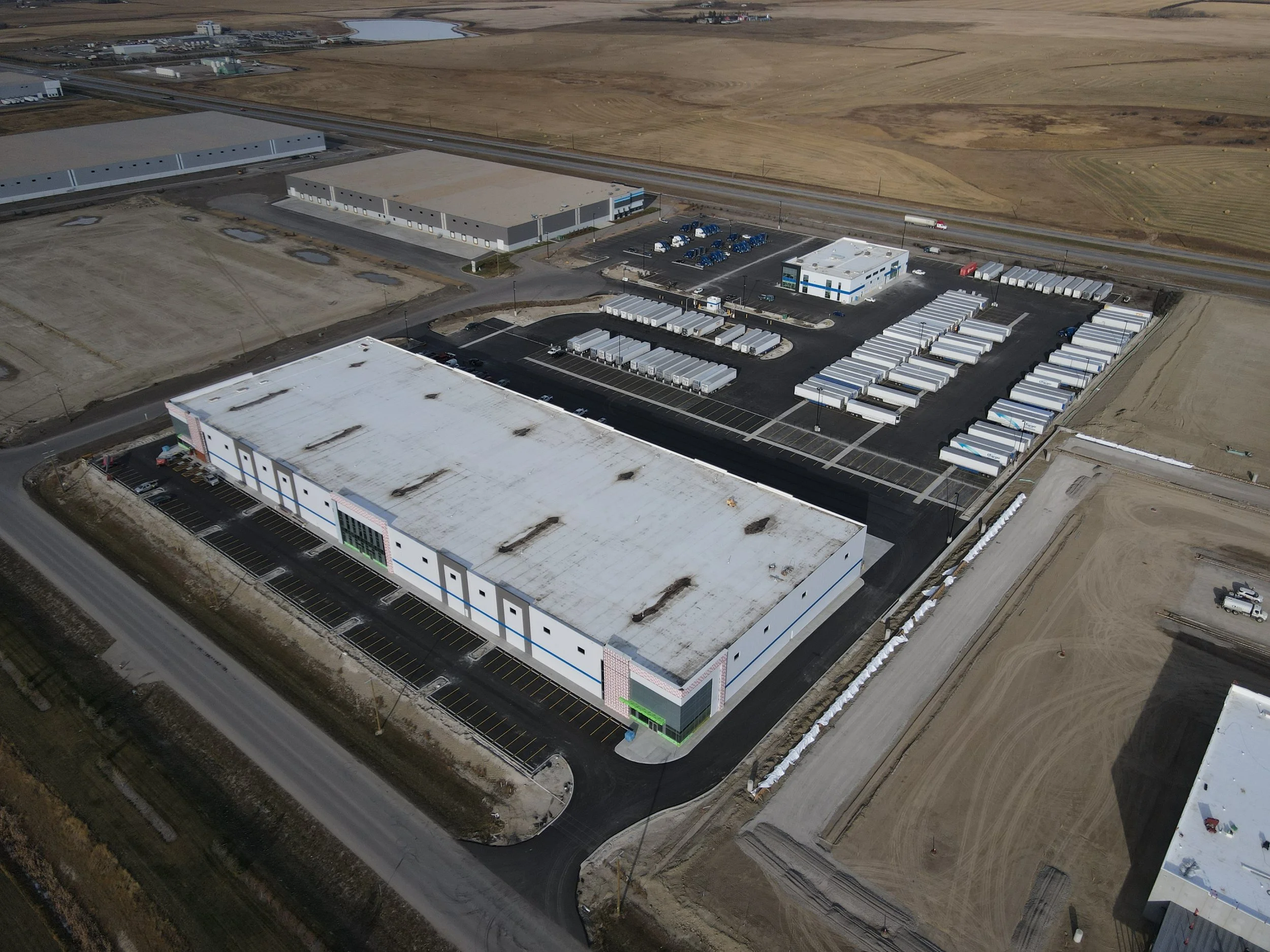 Aerial view of an industrial complex with large warehouse and parking lot, with trailers and trucks parked, surrounded by undeveloped land.