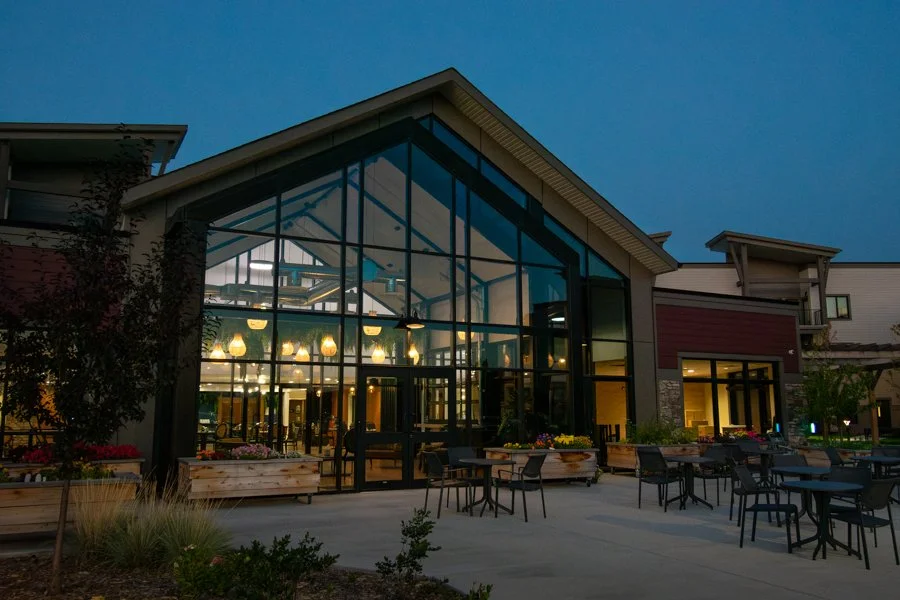 Exterior of a modern building with large glass windows, outdoor seating with tables and chairs, and some flower planters, during evening hours.