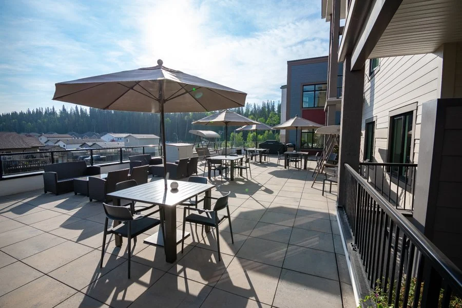 Balcony outdoor seating area with tables, chairs, large umbrellas, and a view of neighboring houses and trees.