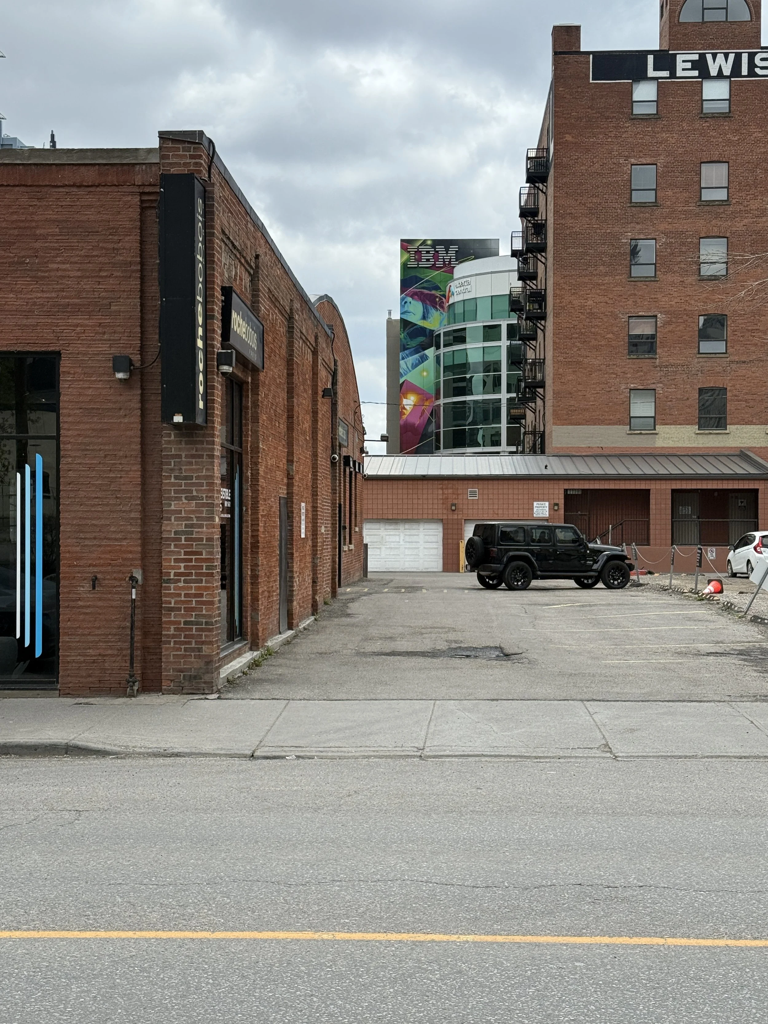 City parking lot with an adjacent red brick building, a black Jeep, and a white car. In the background, modern glass buildings and a colorful billboard with the IBM logo are visible, under a cloudy sky.