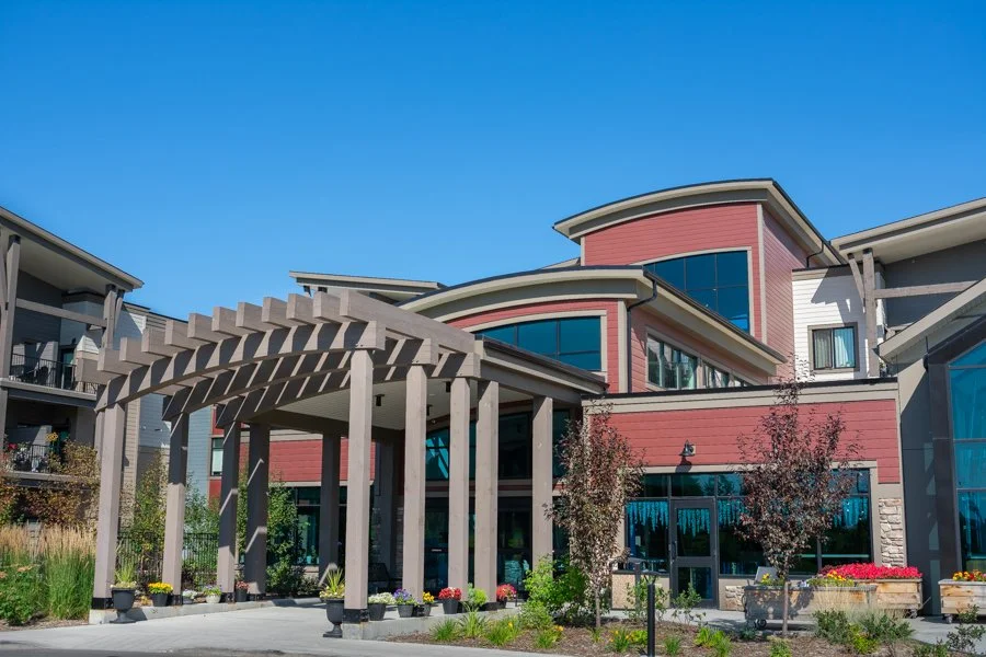 Modern apartment building with a red facade, large windows, and a wooden pergola entrance, surrounded by landscaped gardens under a clear blue sky.