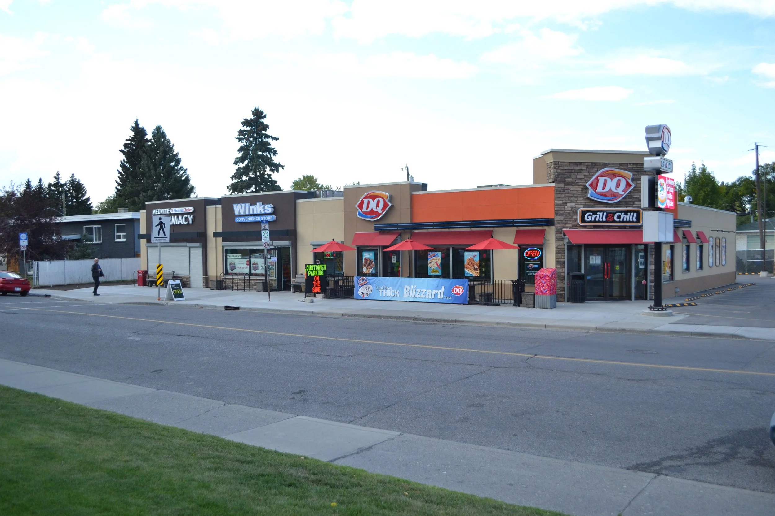 A small commercial shopping strip with stores including Dairy Queen, Winks convenience store, and a pharmacy, located along a city street with trees in the background and a few pedestrians.