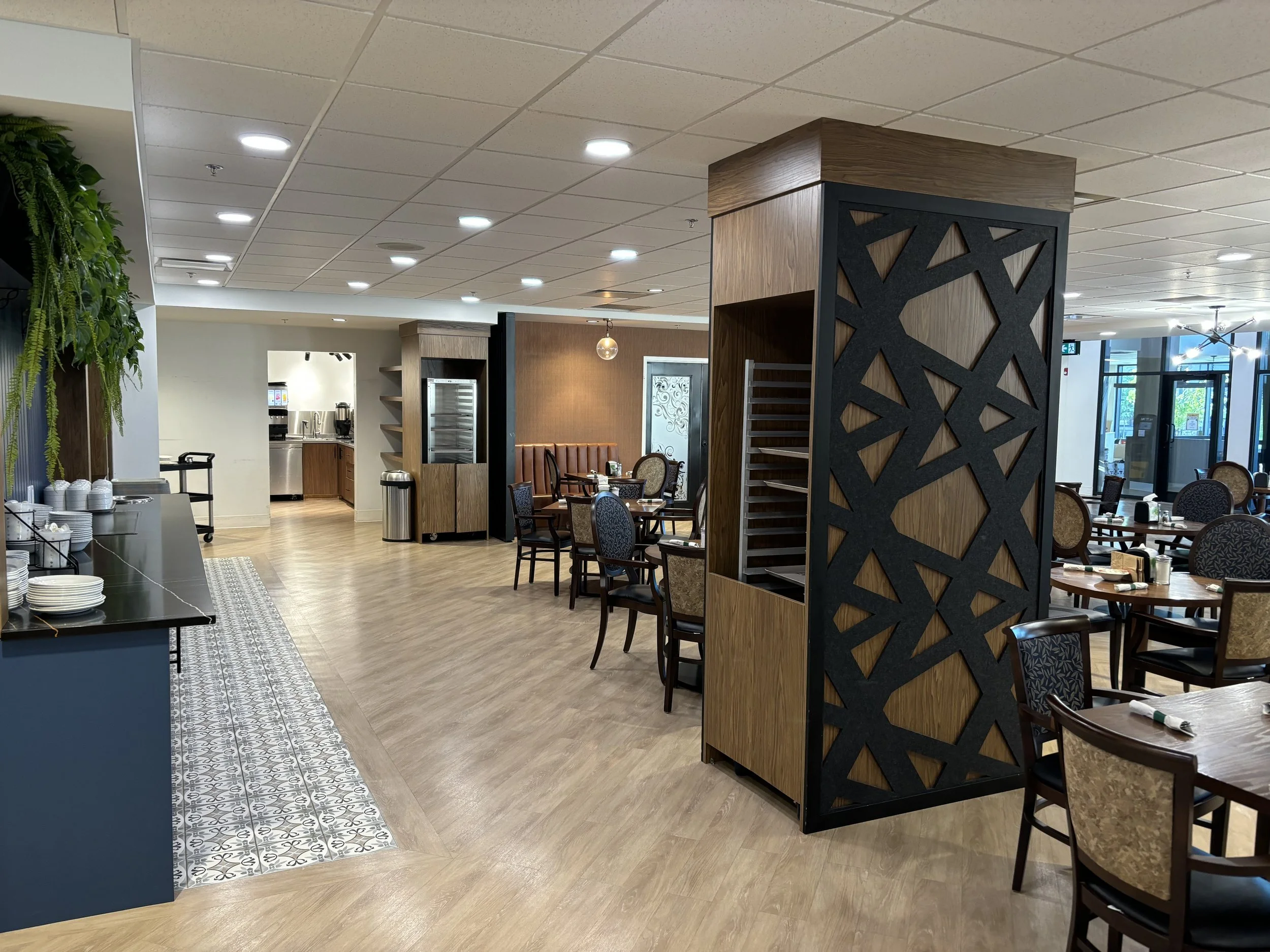 Empty dining area in a restaurant or hotel with wooden tables and chairs, decorative wall panels, a buffet area with plates and cups, and bright lighting.