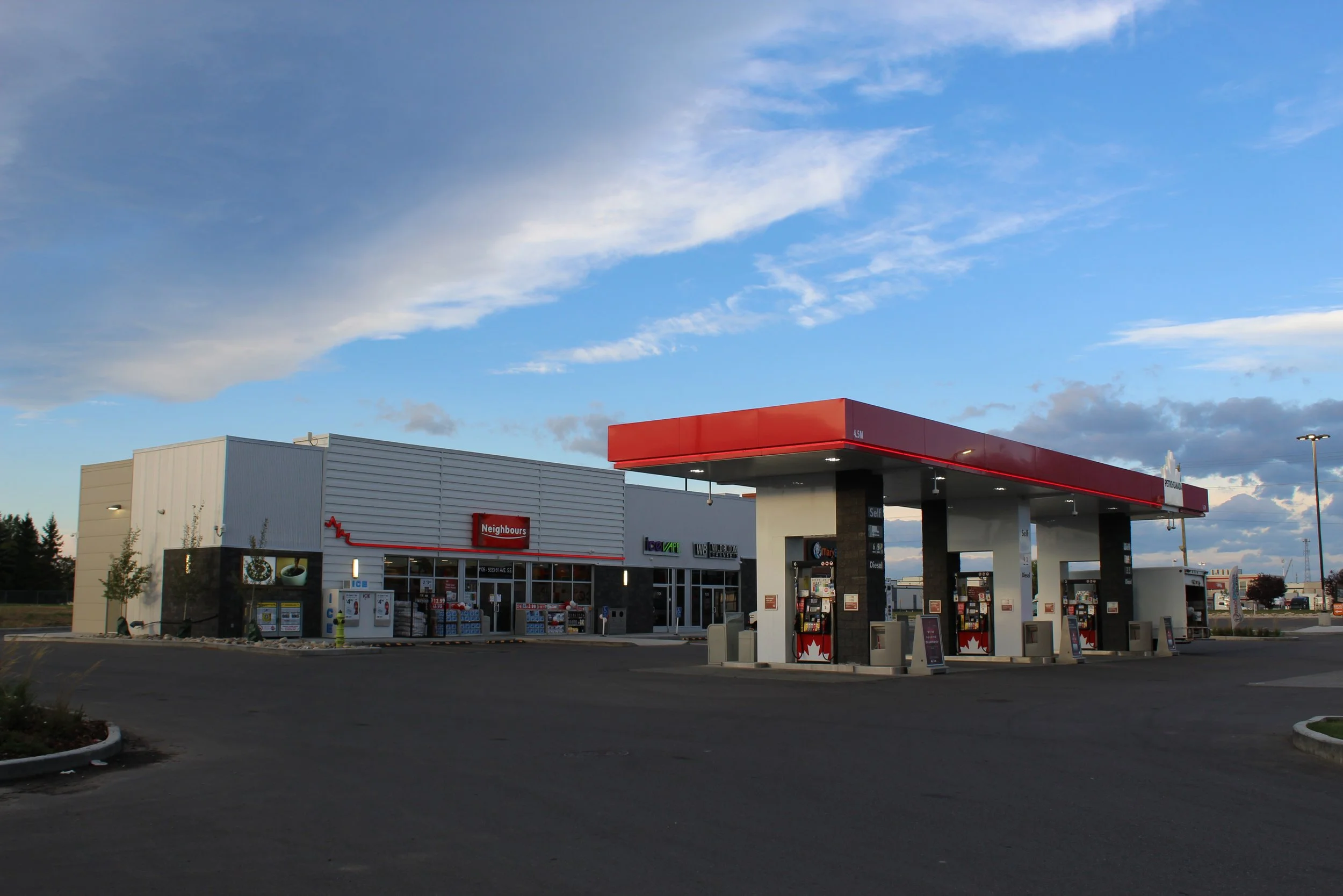 A petrol station with three fuel pumps and a convenience store in the background, under a partly cloudy sky.