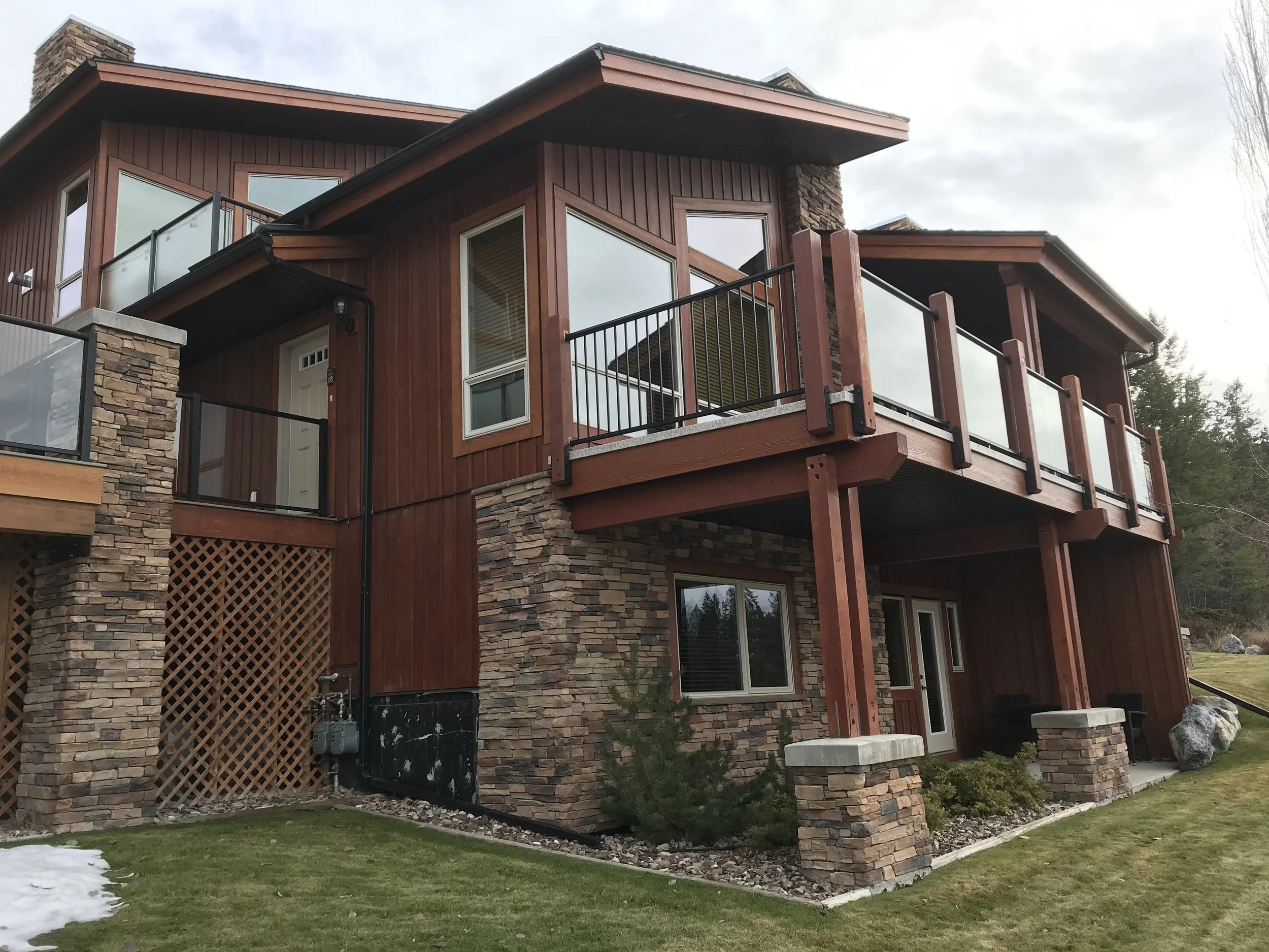 A multi-story house with a stone and wood exterior, featuring large windows and multiple balconies, situated in a grassy yard with some snow and trees in the background.