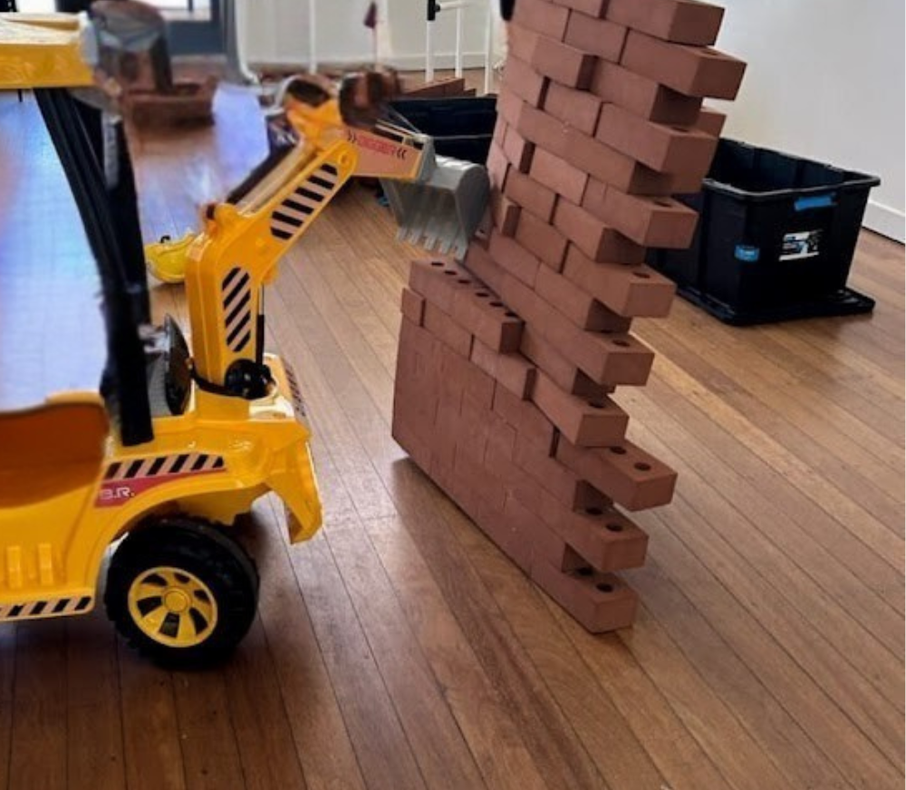 Stacked red bricks arranged in a leaning wall next to a yellow toy construction vehicle on a wooden floor.