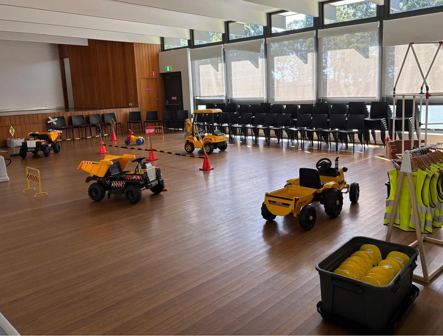 Indoor children's play area with toy construction vehicles, cones, and safety signs, adjacent to a row of black chairs and a clothing rack with reflective vests.