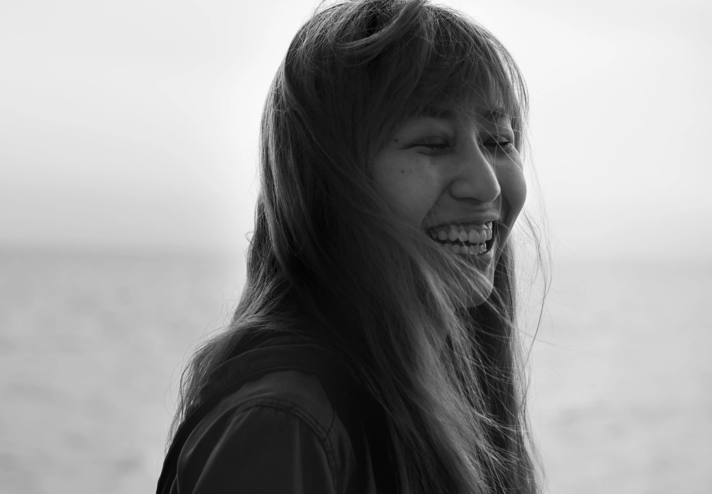 Black and white photo of a woman with long hair, smiling and looking to the side, with a blurred background behind her.