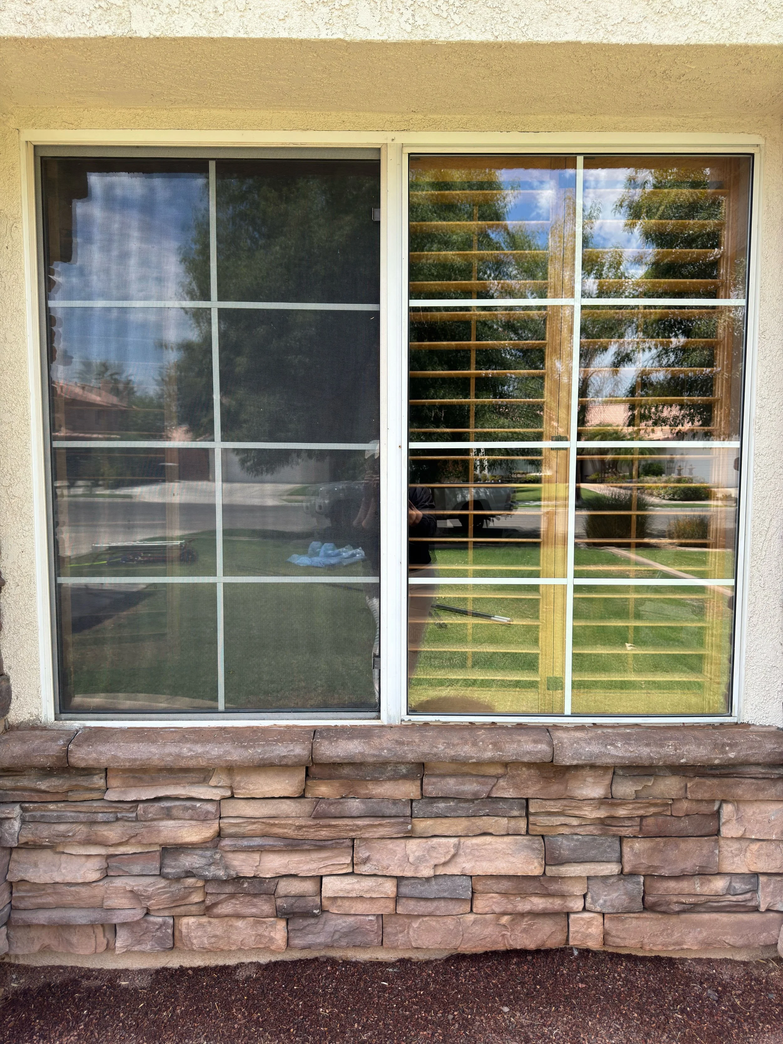 A sliding glass window with a screen door on the left and wooden blinds behind the glass on the right, reflecting the yard and trees outside.