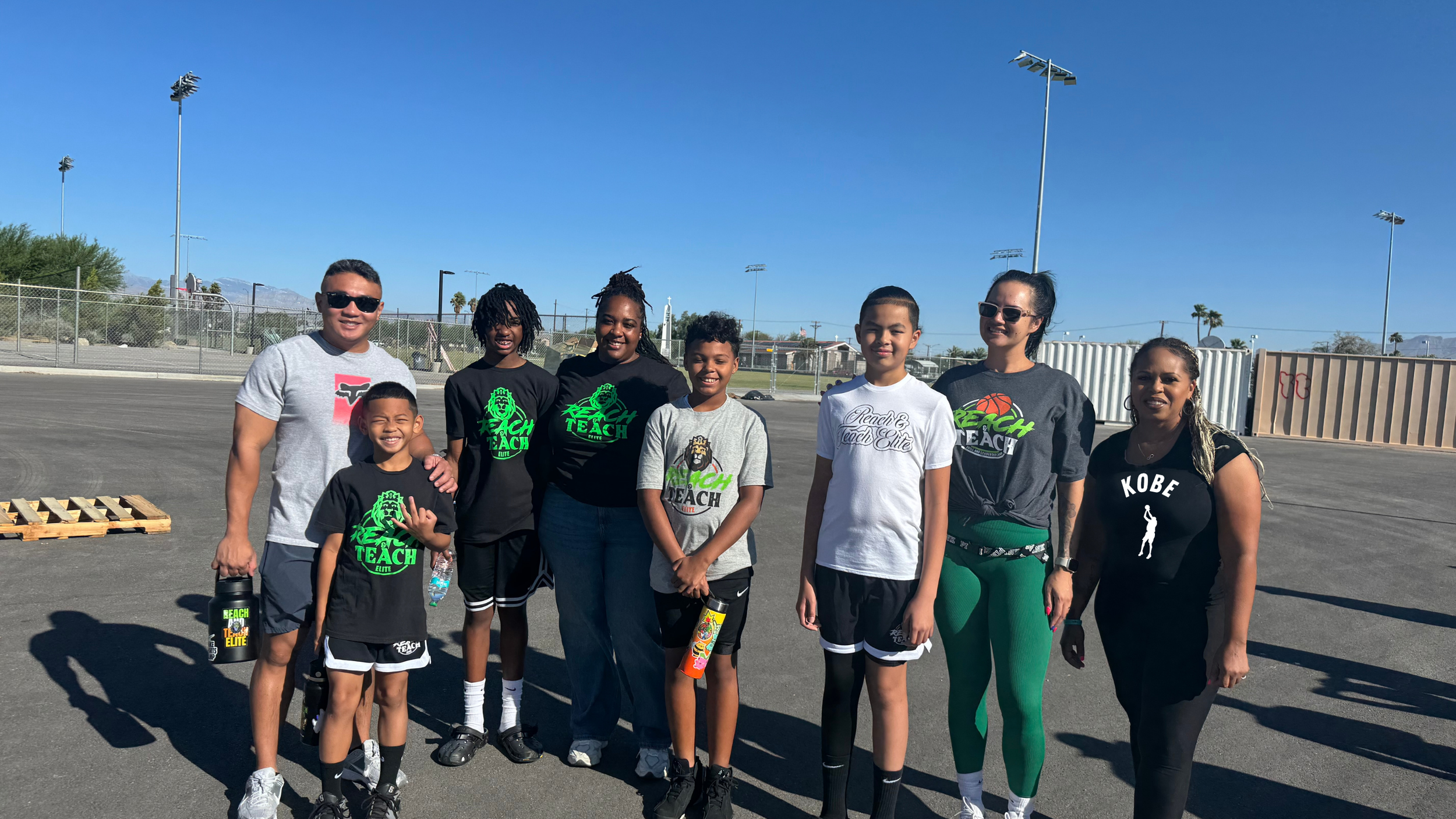 Group of eight people, including children and adults, standing outdoors on a sports field under a clear blue sky, posing for a photo.