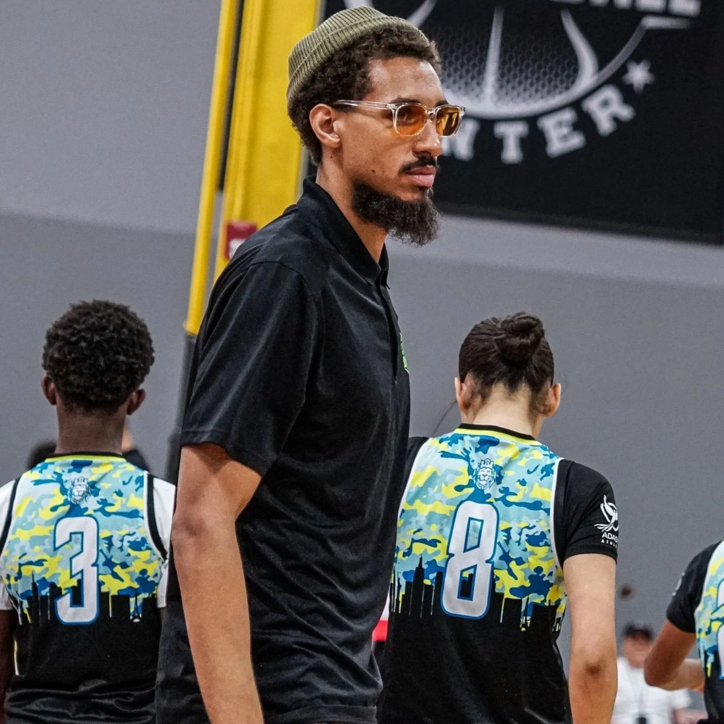 A man with a beard, glasses, and a beanie standing in front of a group of basketball players in jerseys with the numbers 3 and 8.