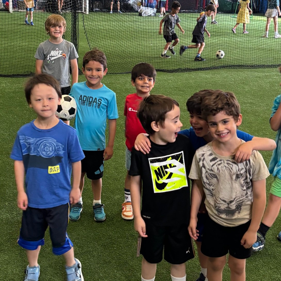 Group of young boys smiling and posing together inside an indoor soccer facility with a netted area in the background where children are playing soccer.