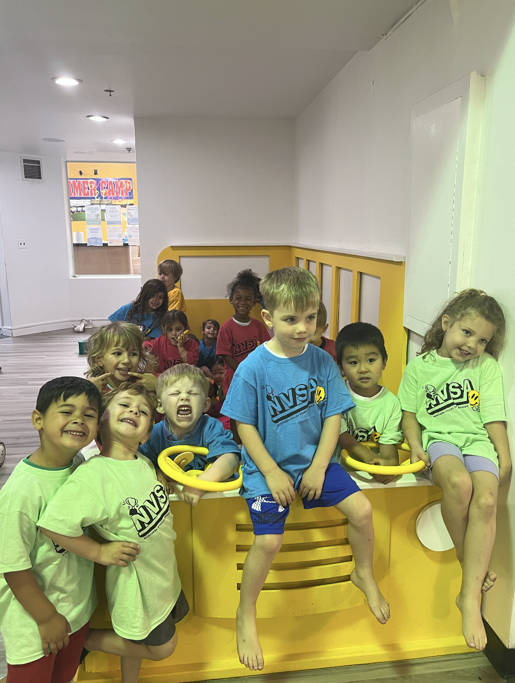 Group of children sitting and standing around a yellow indoor play structure, smiling and making funny faces.