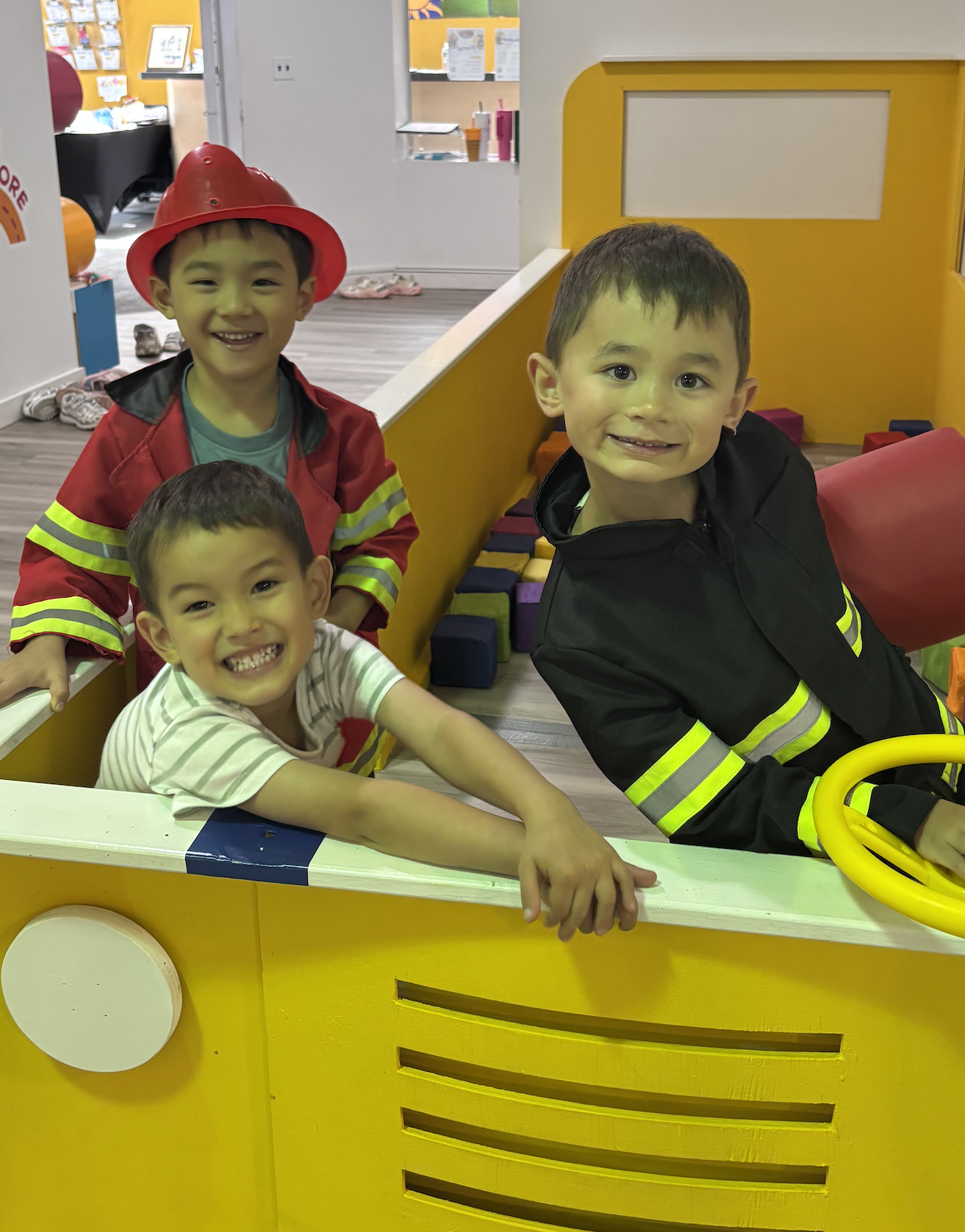 Three young boys playing in a fire truck-themed play area, with two dressed in firefighter costumes.