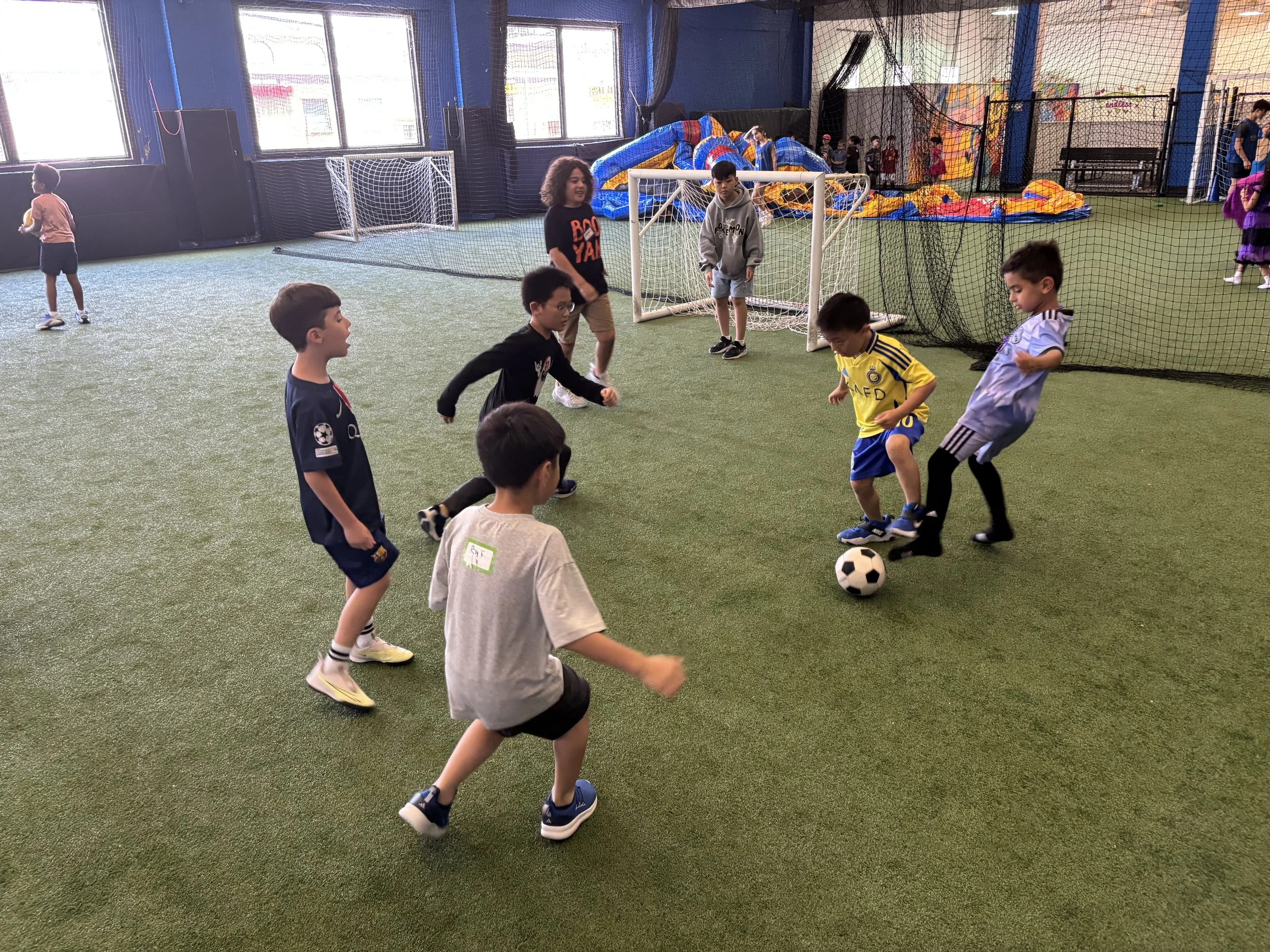 Children playing with a colorful parachute inside an indoor play area with artificial grass, netted ceiling, and sports equipment in the background.