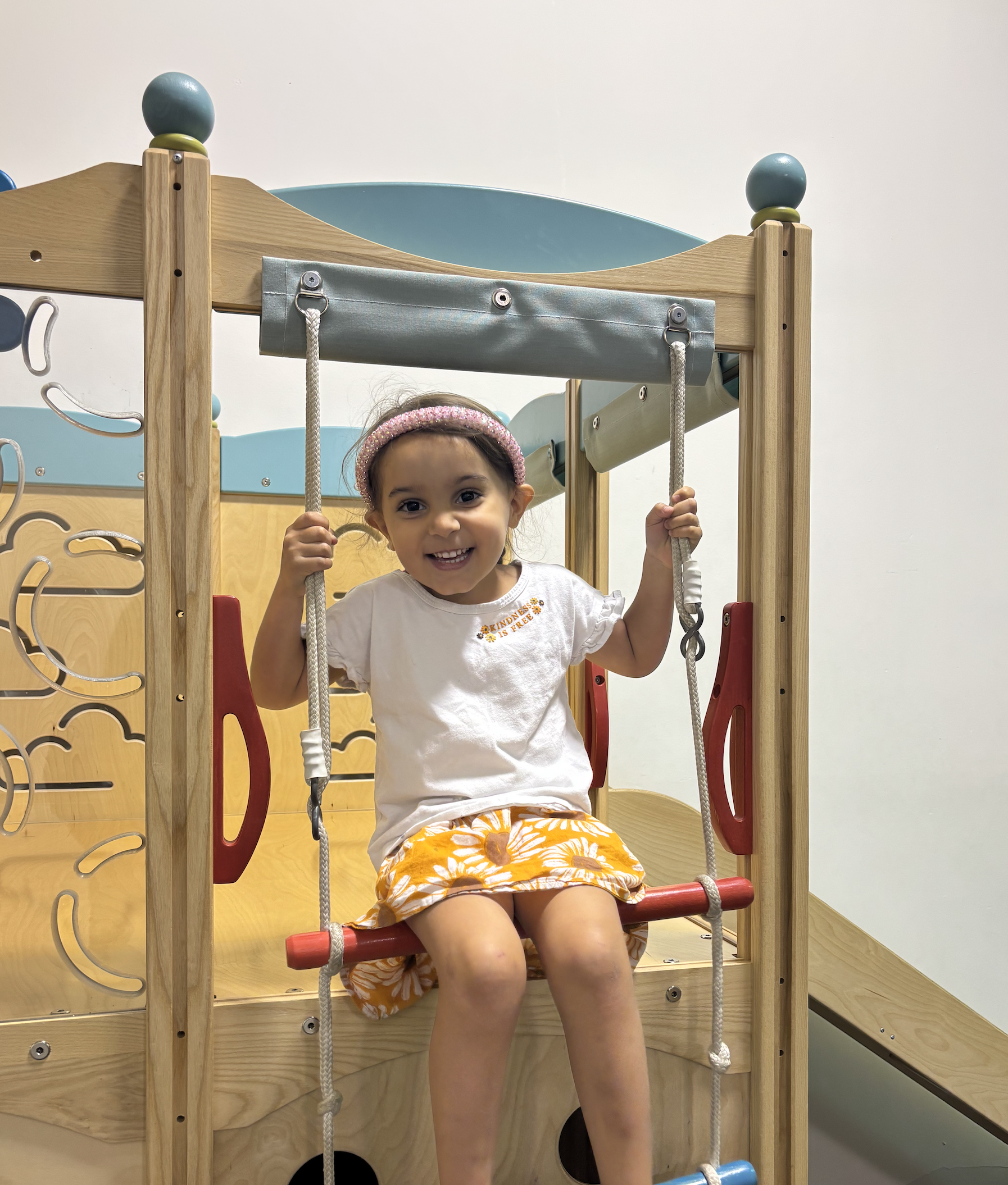 A young girl smiling while sitting on a red swing at a wooden indoor play structure.