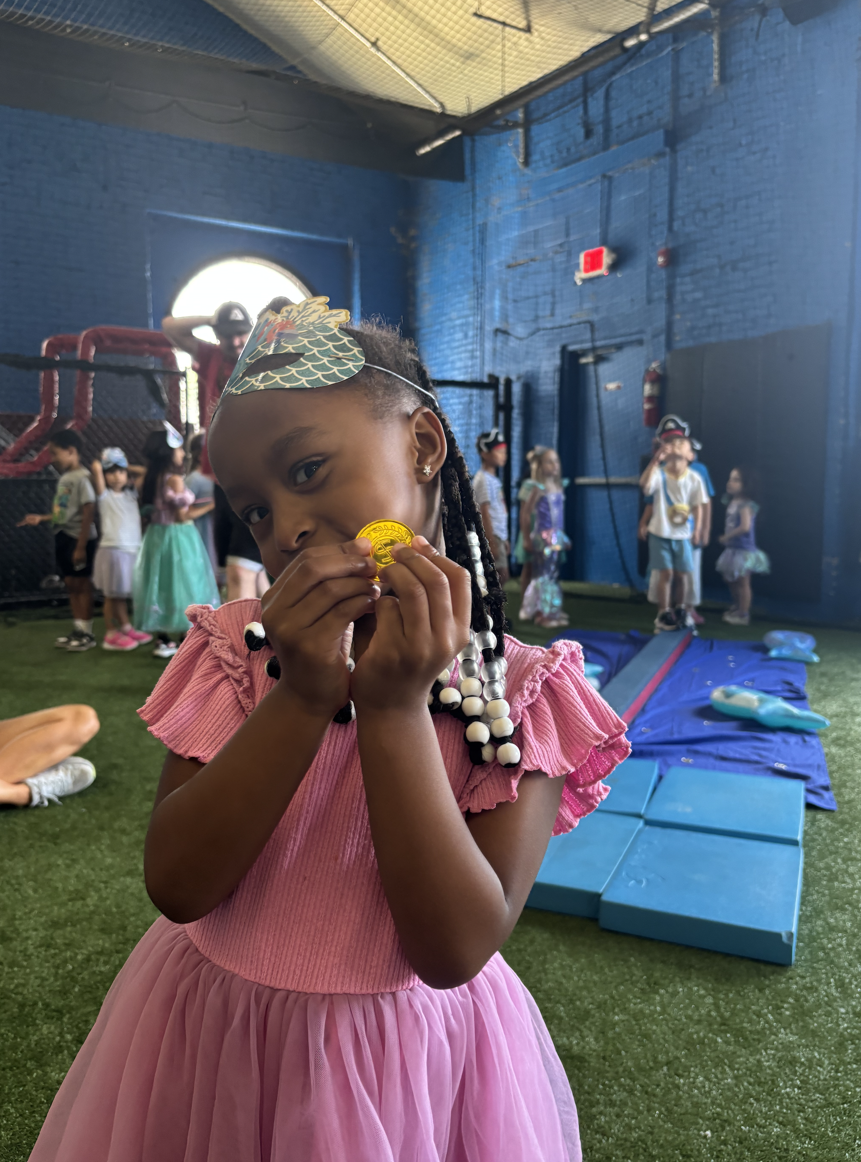 A young girl in a pink dress with beaded hair holds a gold chocolate coin close to her mouth at an indoor party with other children in costumes in the background.