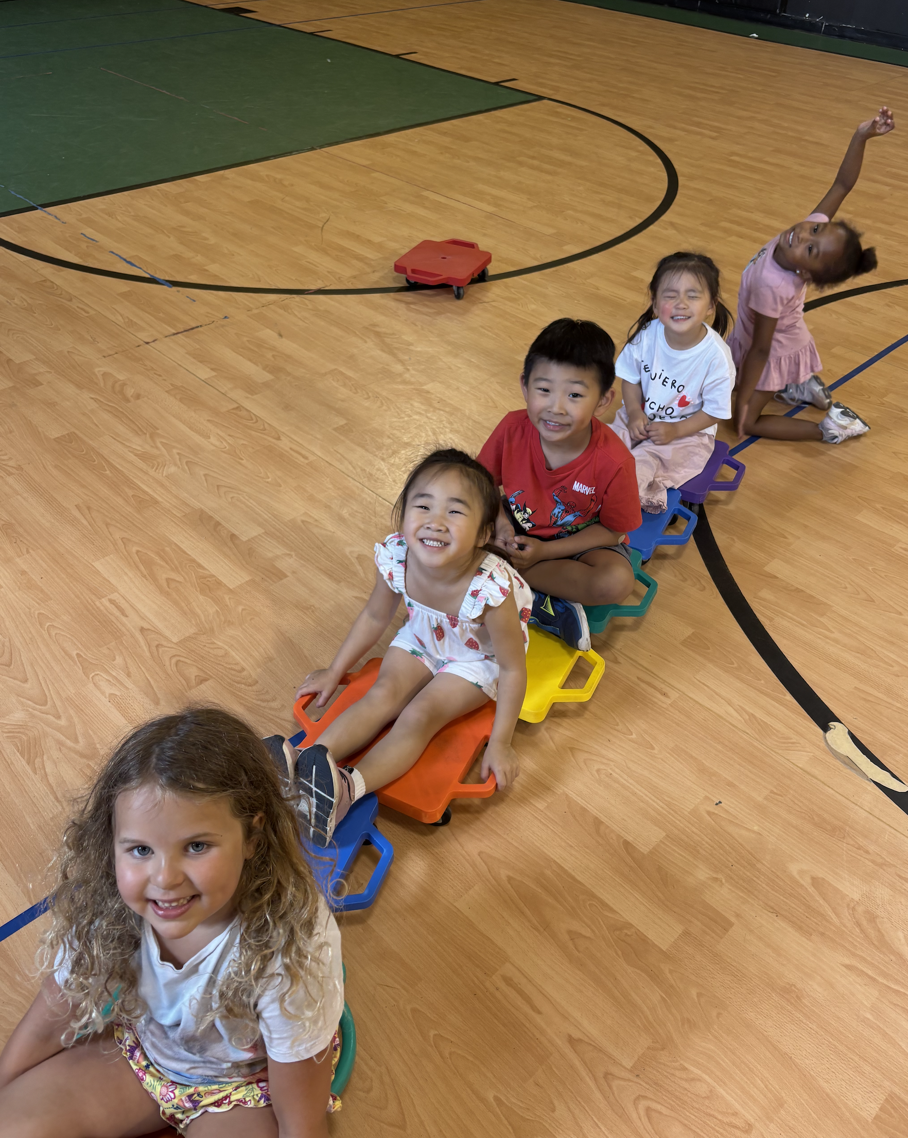 A group of six children sitting and lying on a colorful plastic train on a gymnasium floor, smiling and having fun.