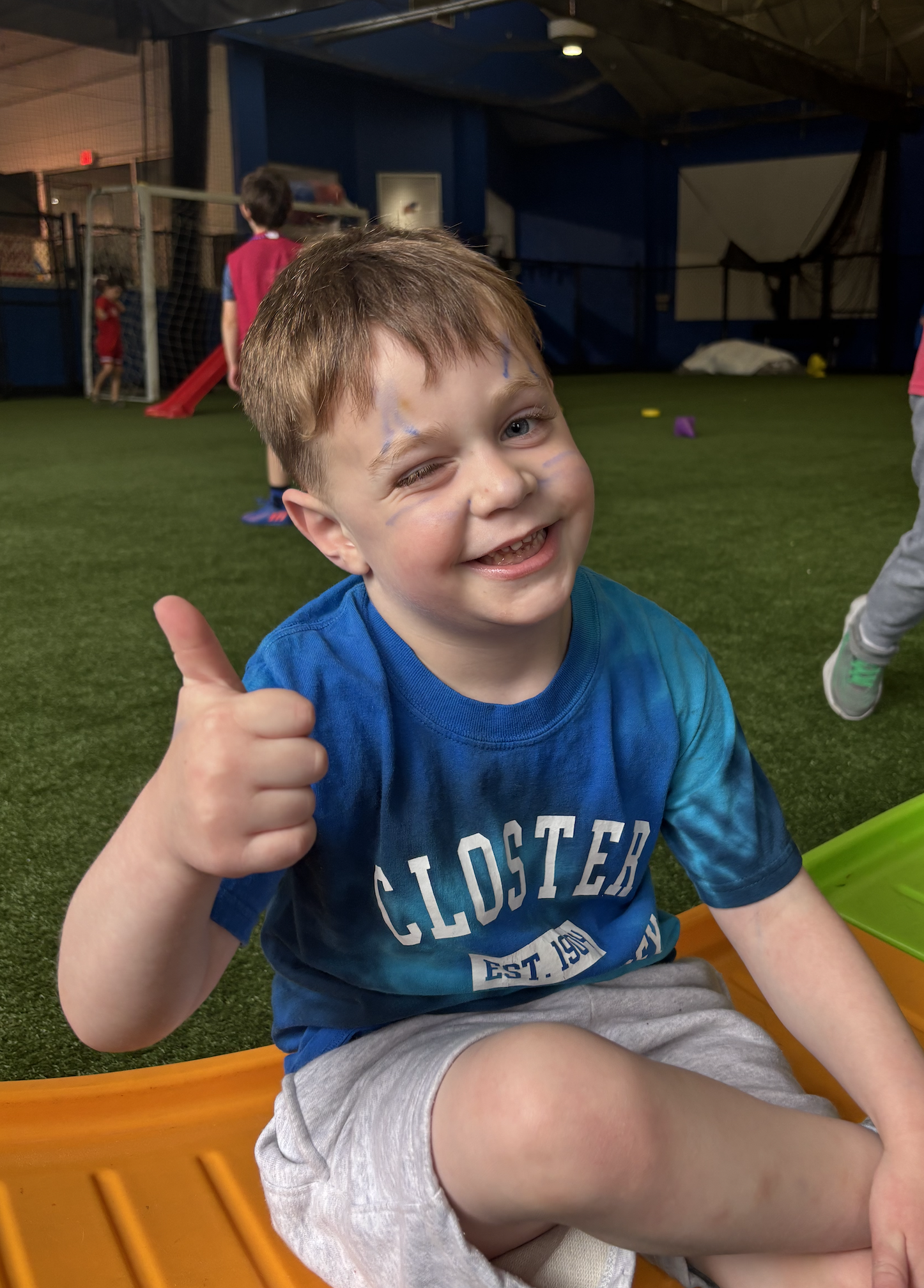 A young boy with face paint smiling and giving a thumbs-up inside an indoor play area with artificial turf.