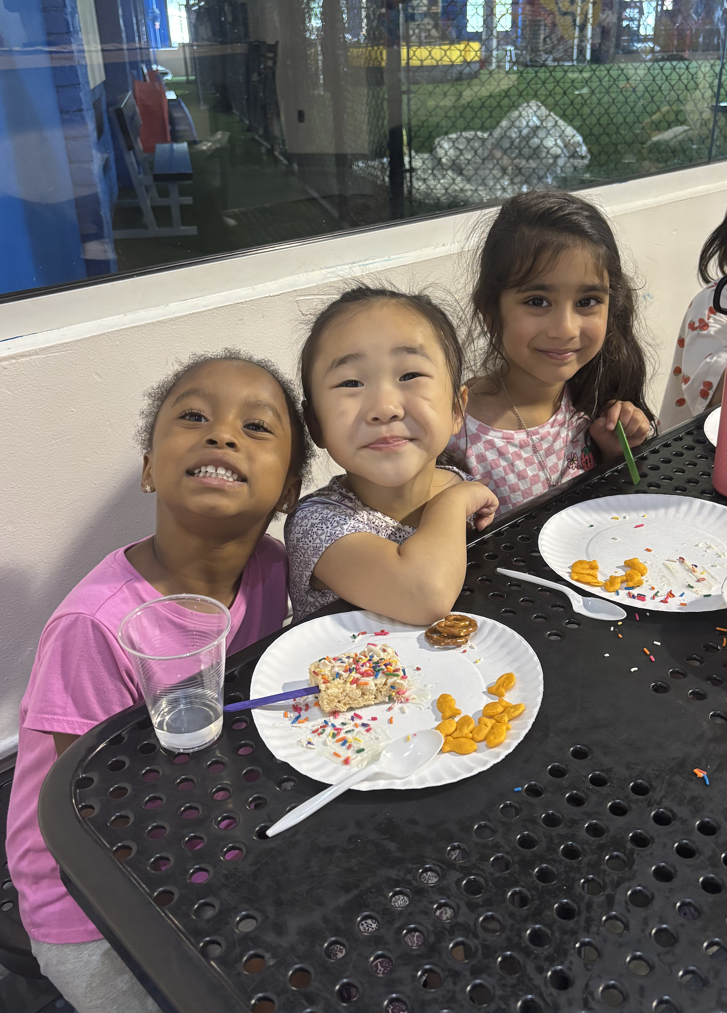 Three young girls sitting at a black metal picnic table during a birthday party, smiling at the camera. Plates with cake and snacks, a glass of water, and birthday decorations are on the table. Behind them is a window showing an outdoor playground.