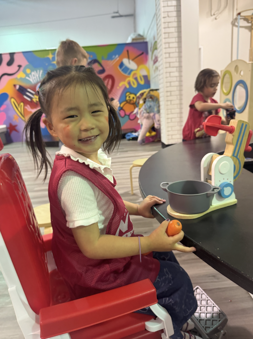 Young girl smiling while playing with toy kitchen set in a colorful playroom, holding a plastic carrot, with other children and vibrant wall art in the background.