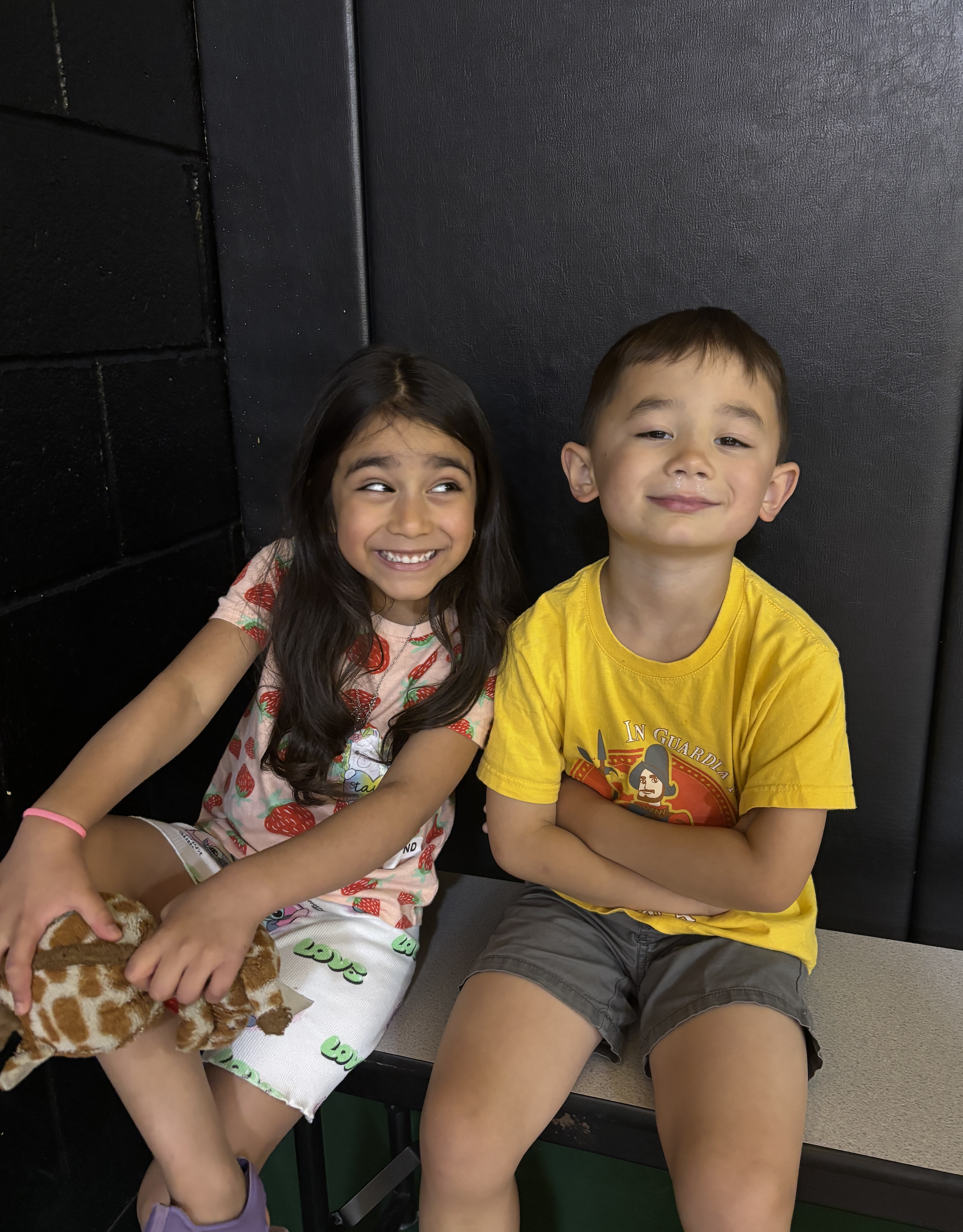 A young girl and boy sitting on a bench against a black background. The girl is smiling and holding a stuffed giraffe, wearing a strawberry-patterned shirt and white skirt with green writing. The boy has his arms crossed, wearing a yellow T-shirt wit