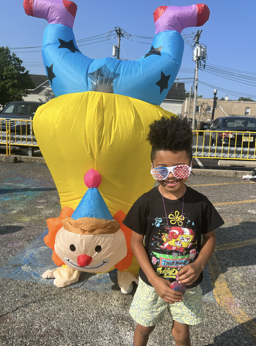 A young boy wearing patriotic sunglasses and a black T-shirt stands in front of colorful Halloween-themed balloons in a parking lot.
