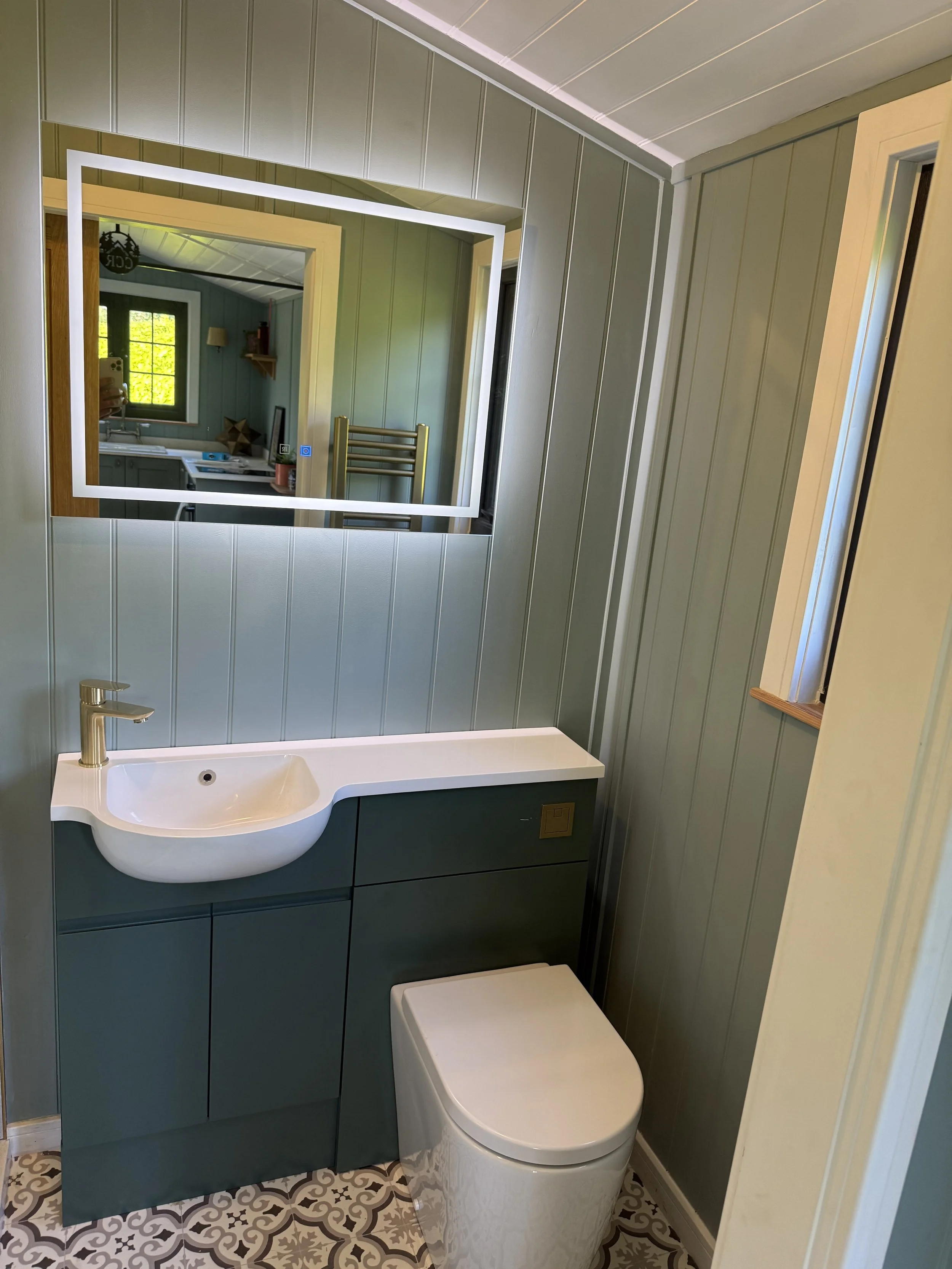 Small bathroom with patterned tile floor, green paneling walls, a wall-mounted mirror with LED lights, a white sink with silver faucet, a closed toilet, and a window showing greenery outside.