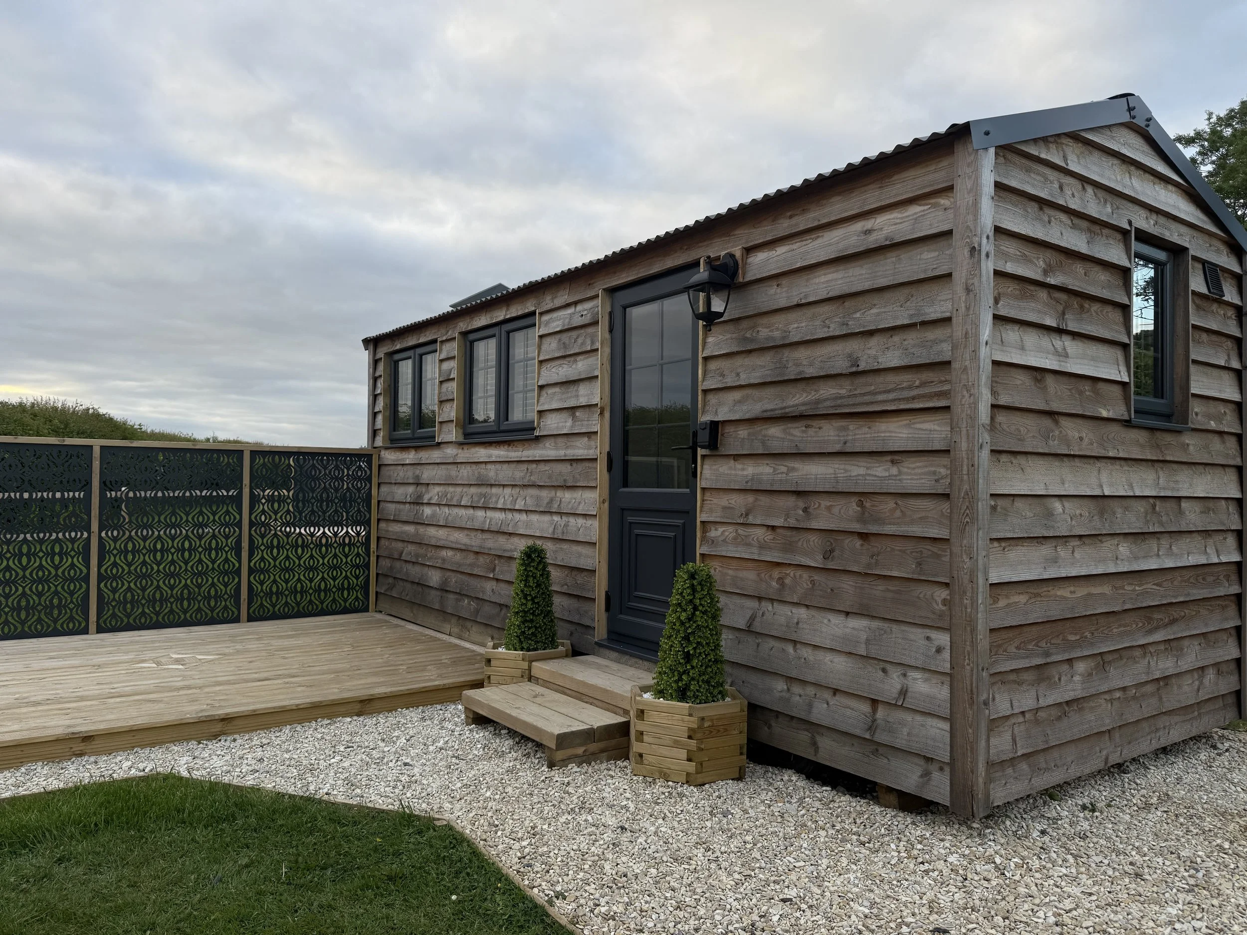 A small wooden house with horizontal wooden siding, black window and door frames, and a gray metal roof, situated in a yard with a deck, two potted bushes, a gravel area, and a green lawn, under a cloudy sky.